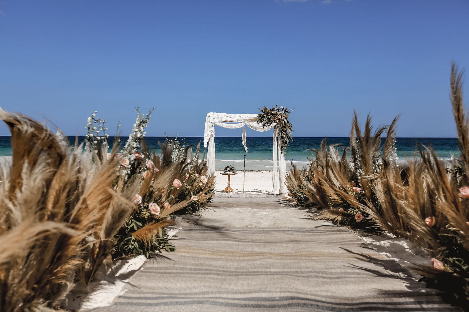 Beach wedding aisle decorated with dried pampas grass leading to a white arch with ocean views.