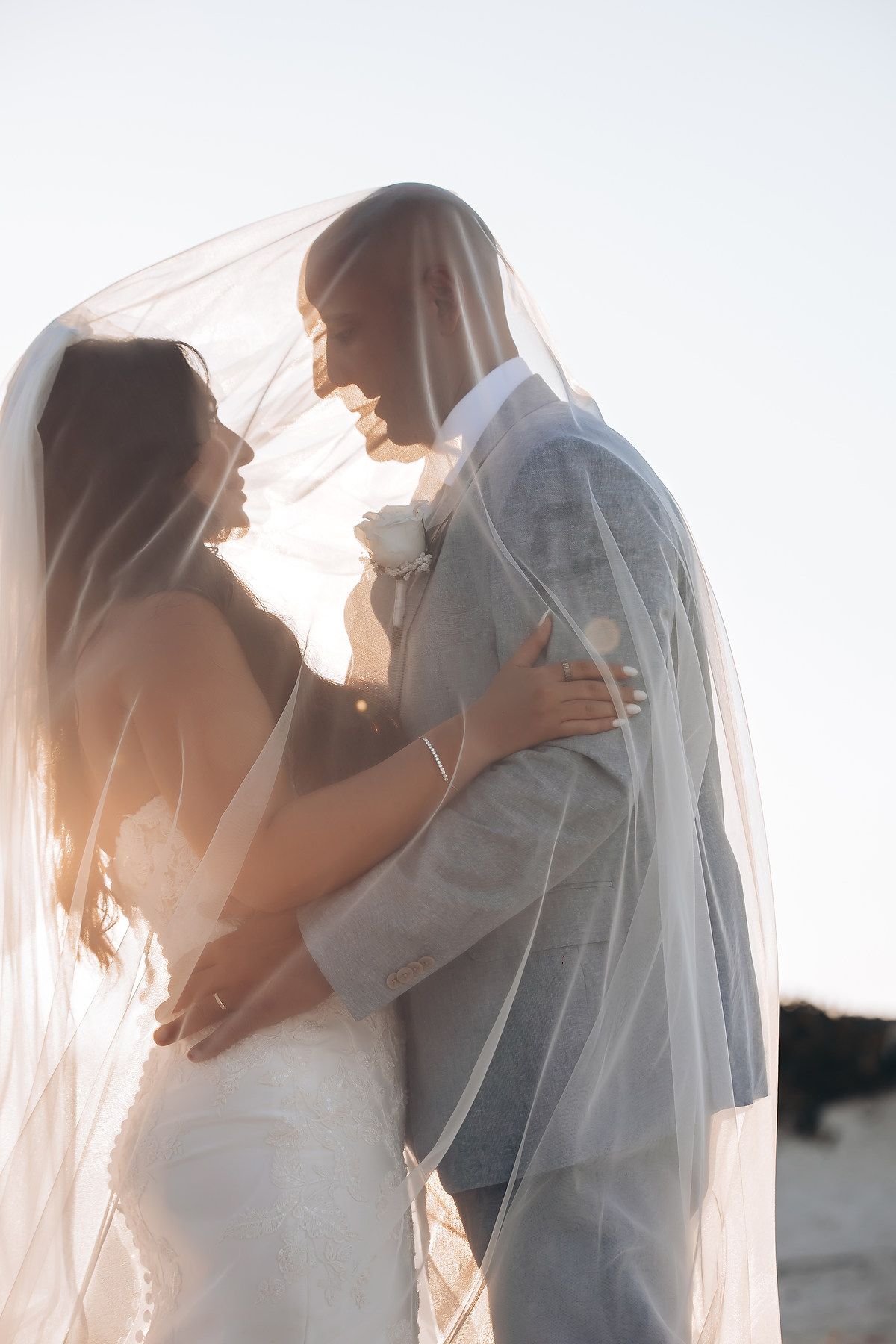 A romantic wedding couple embraces in soft sunlight creating silhouettes through a delicate veil.