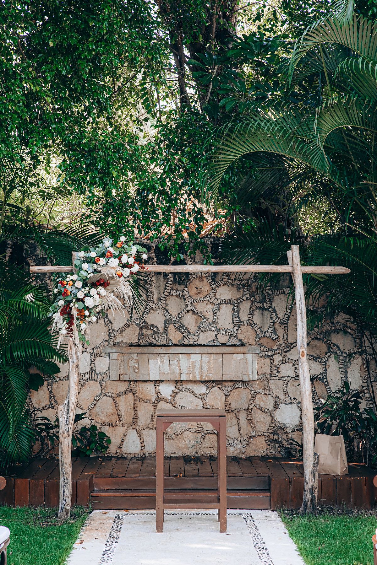 Wooden wedding arch decorated with white and pink flowers against a stone wall in a tropical garden setting.