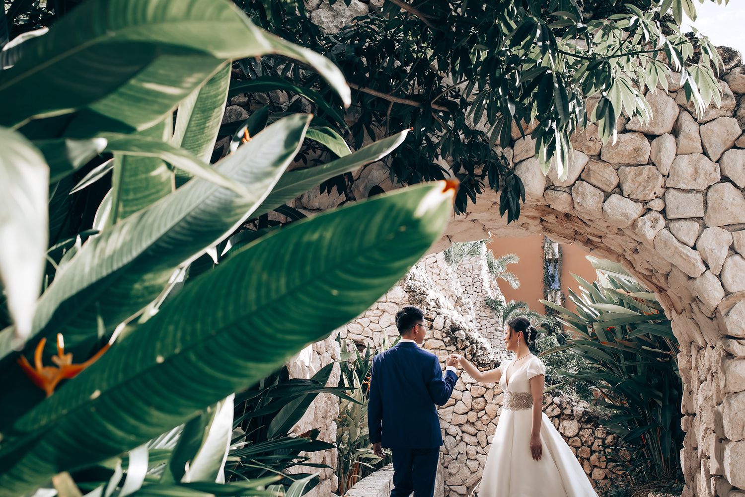 Wedding couple photographed through tropical green leaves against stone wall backdrop.