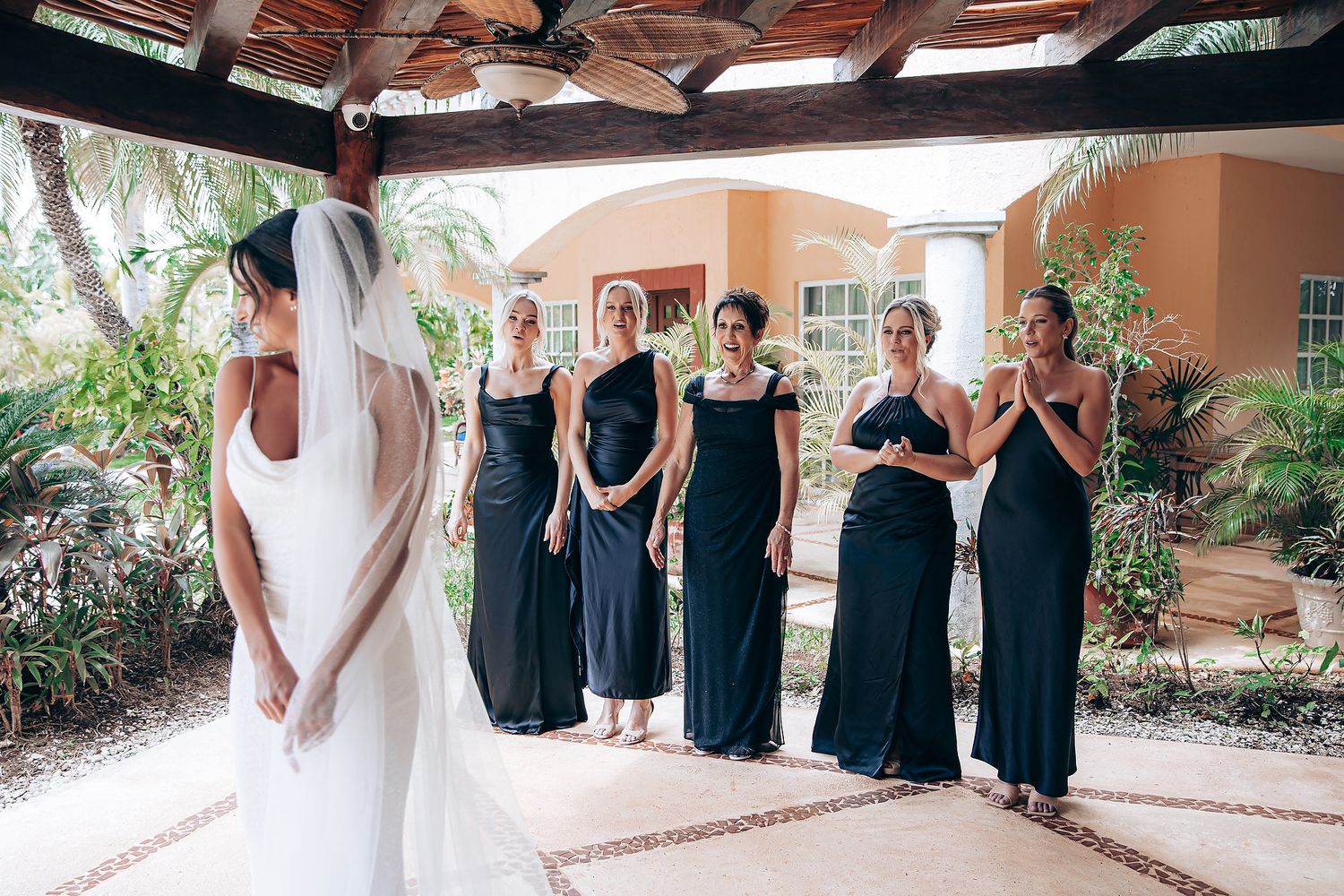 Wedding party in black dresses gathered under a covered terrace during pre-ceremony preparations.