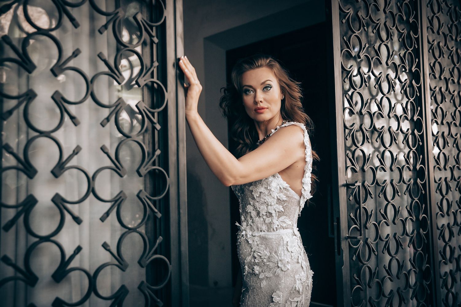 Bride in white lace dress stands in dramatic lighting against decorative iron gate backdrop.