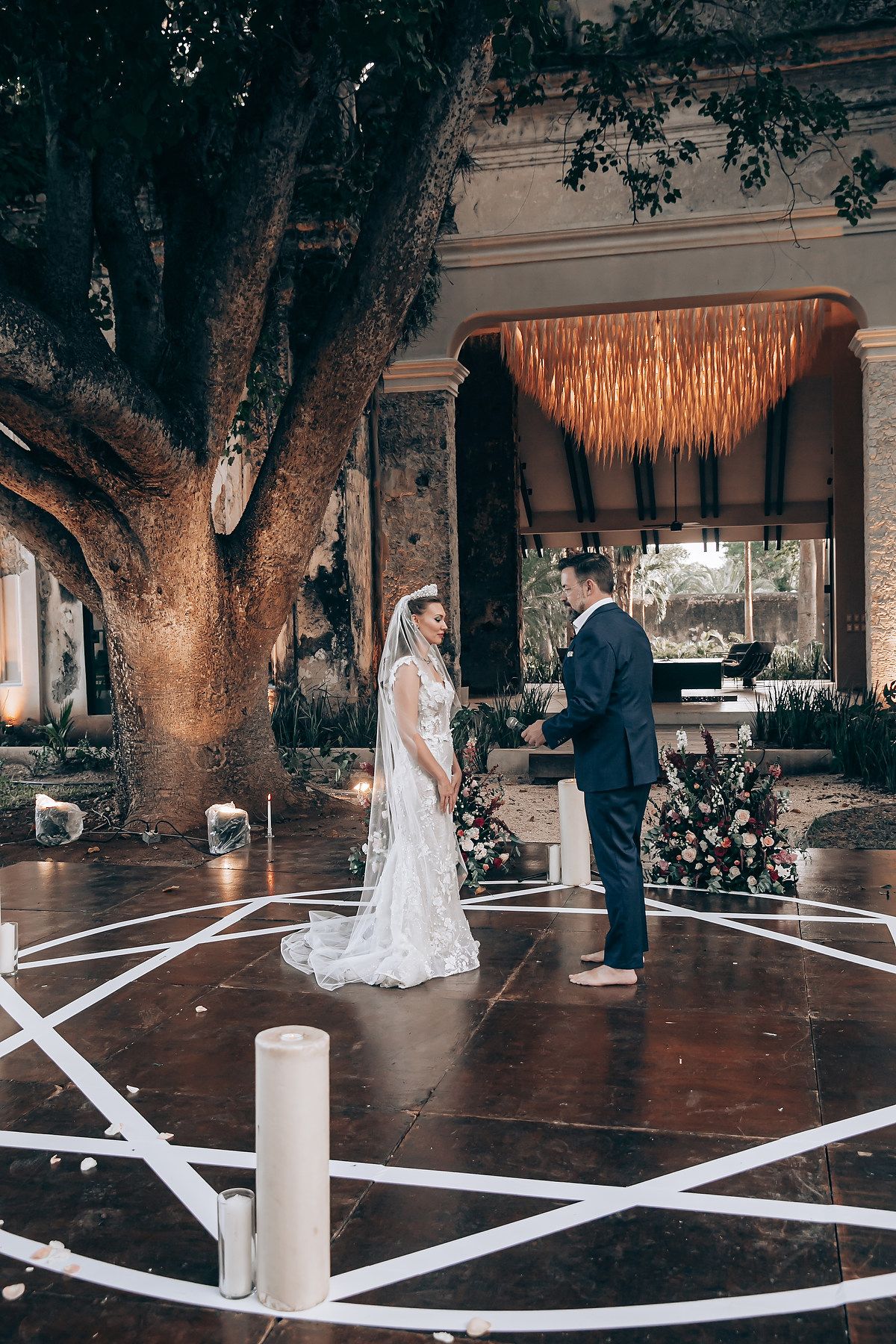 A wedding ceremony takes place under a grand tree with white geometric floor designs and a bohemian chandelier overhead.