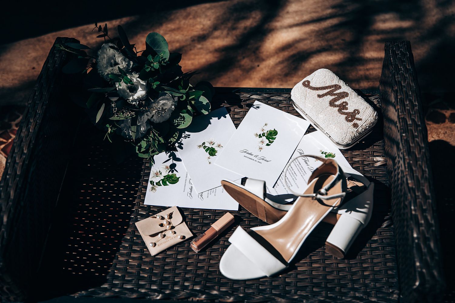 Wedding invitation cards and floral decorations arranged on dark textured surface with elegant white sandals.
