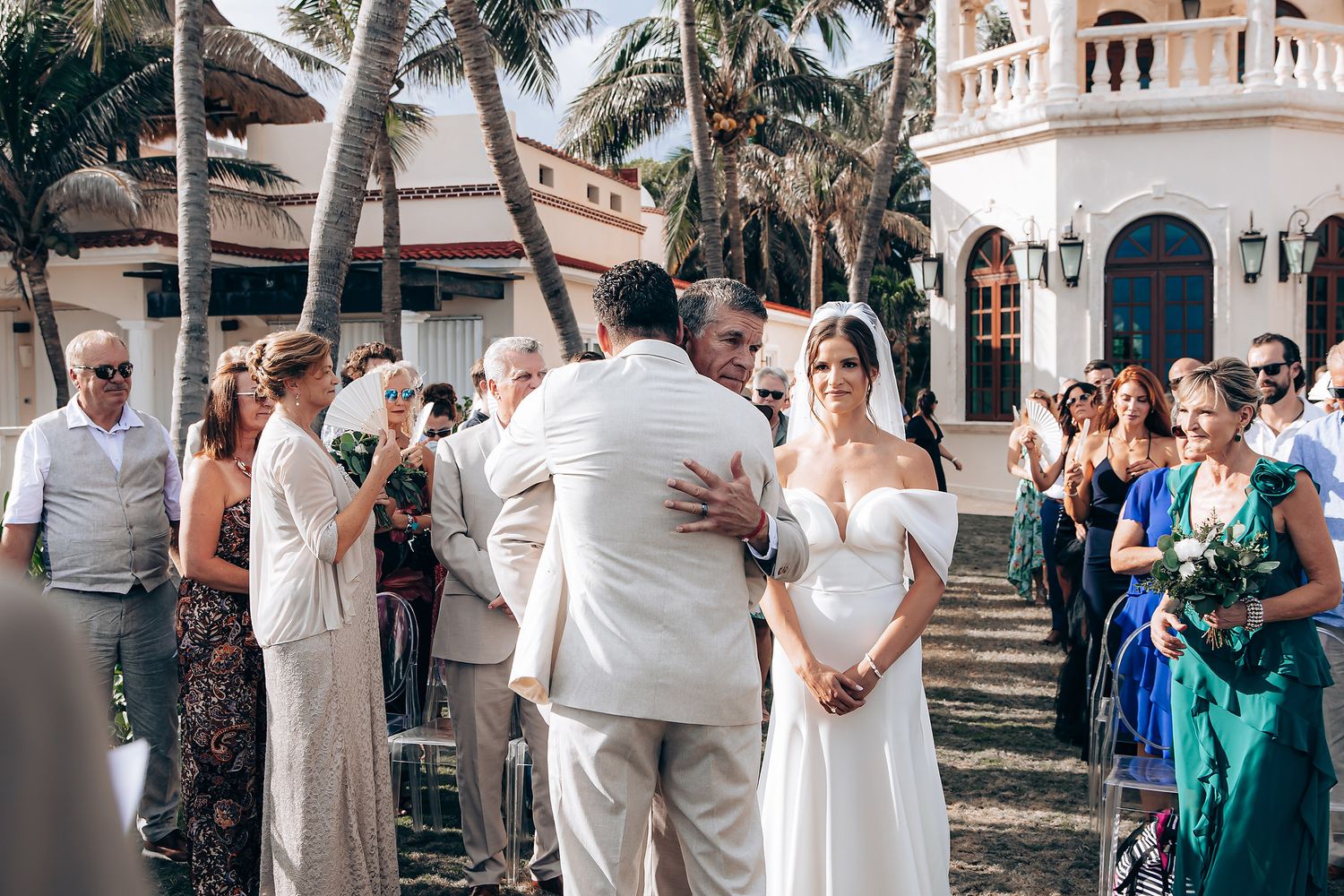 An outdoor wedding ceremony takes place on a tropical property with palm trees and Mediterranean-style architecture.