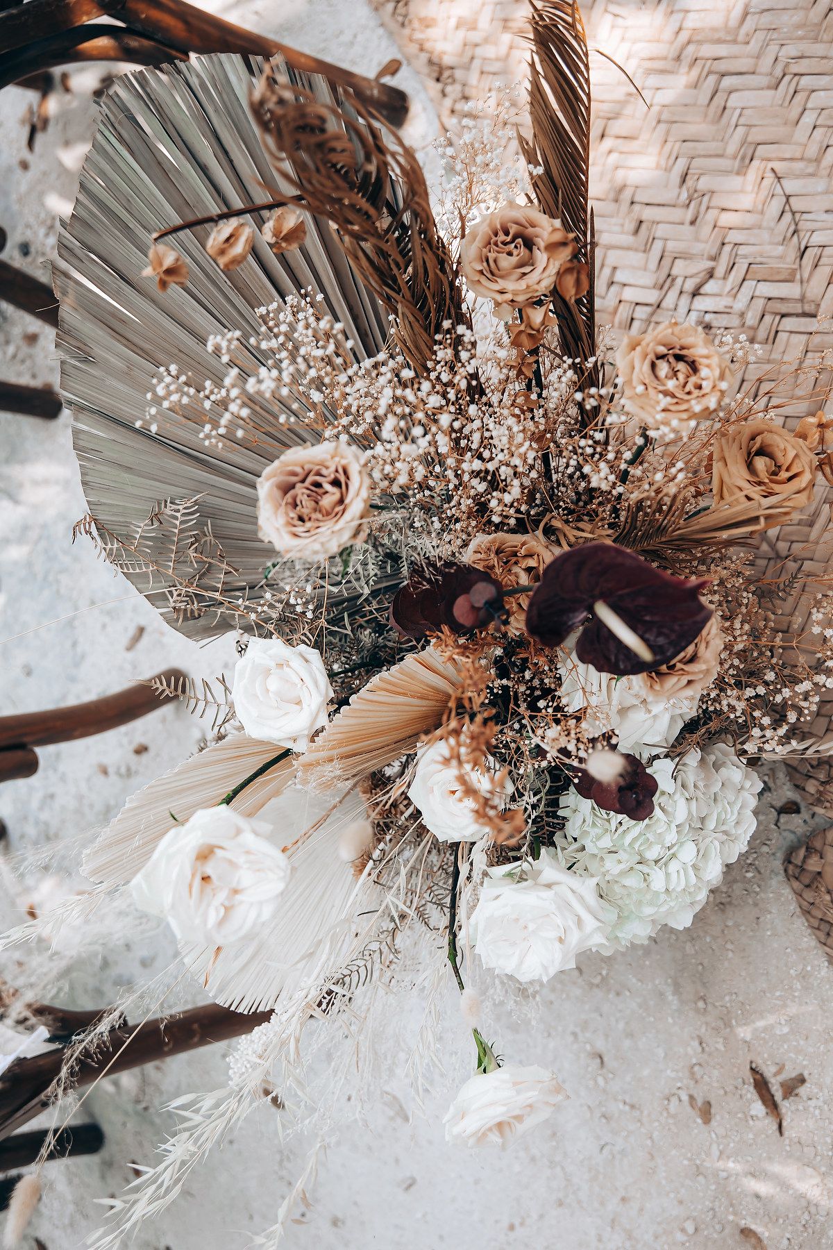 Dried floral arrangement featuring palm fronds, roses and baby's breath in neutral tones against a woven backdrop.