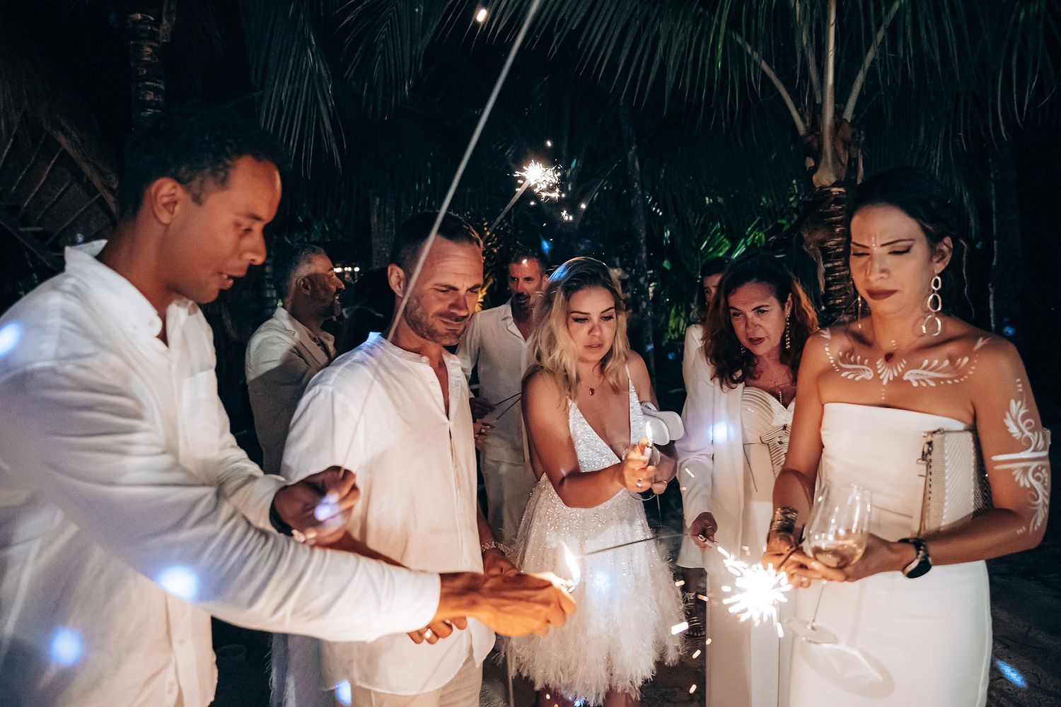 Wedding party in white attire celebrates with sparklers during an outdoor evening reception.