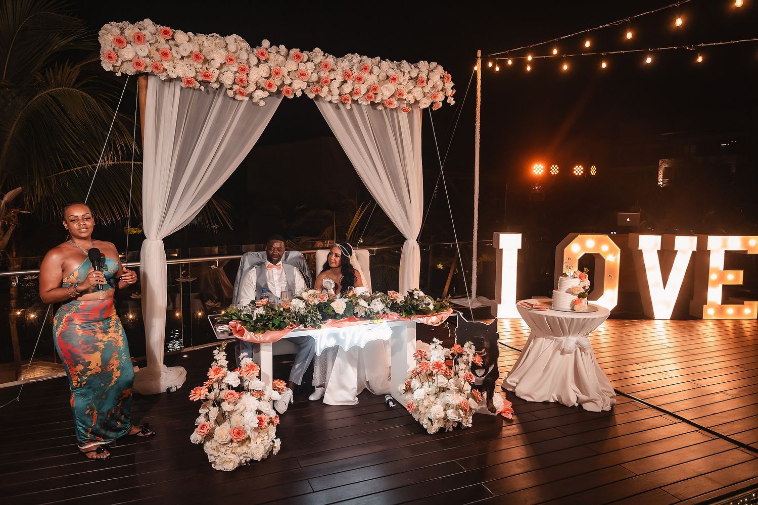 Wedding reception setup with string lights, floral arrangements, and illuminated LOVE sign at night.