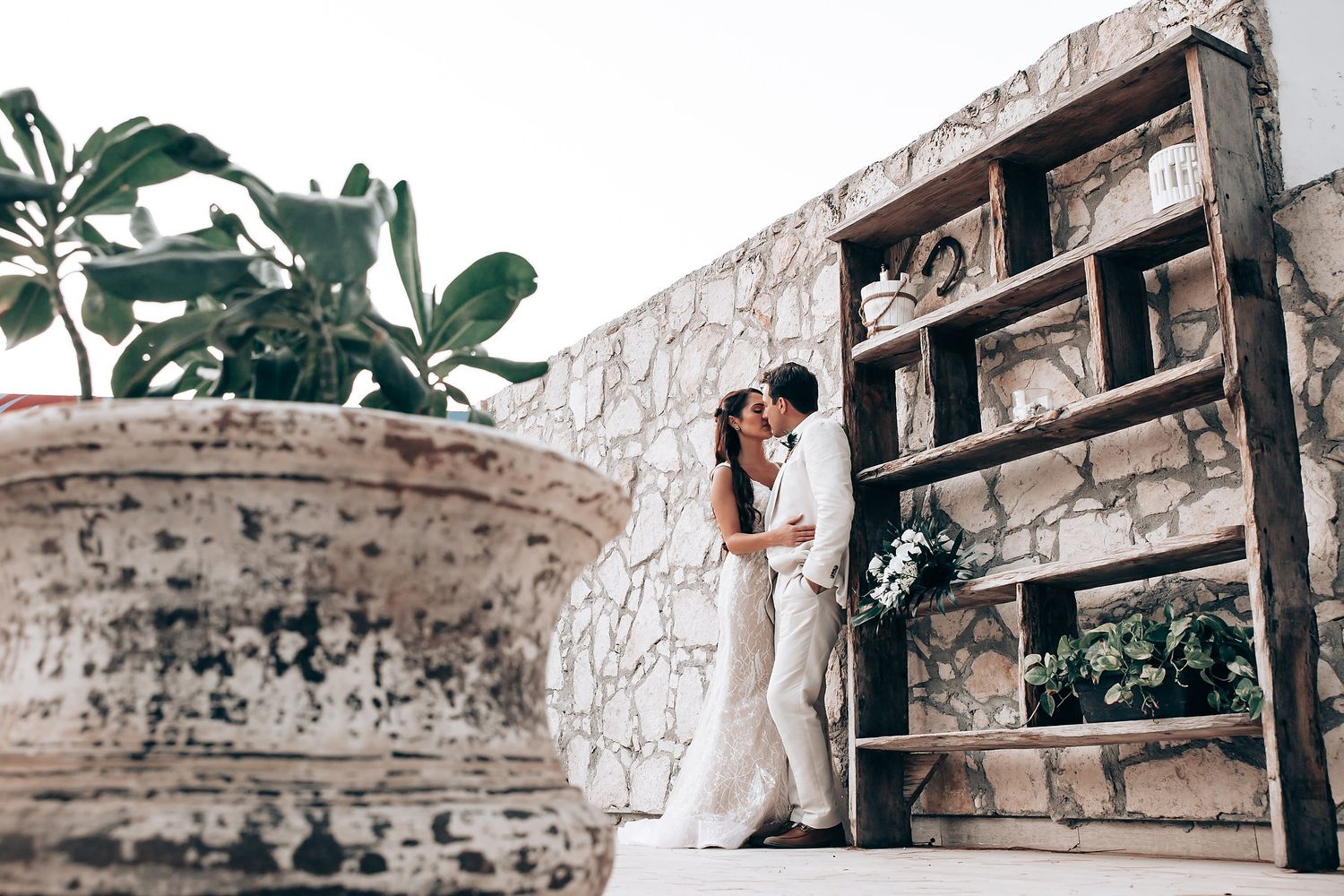 Elegant wedding couple embraces on stone steps next to vintage wooden shelving and potted plants.