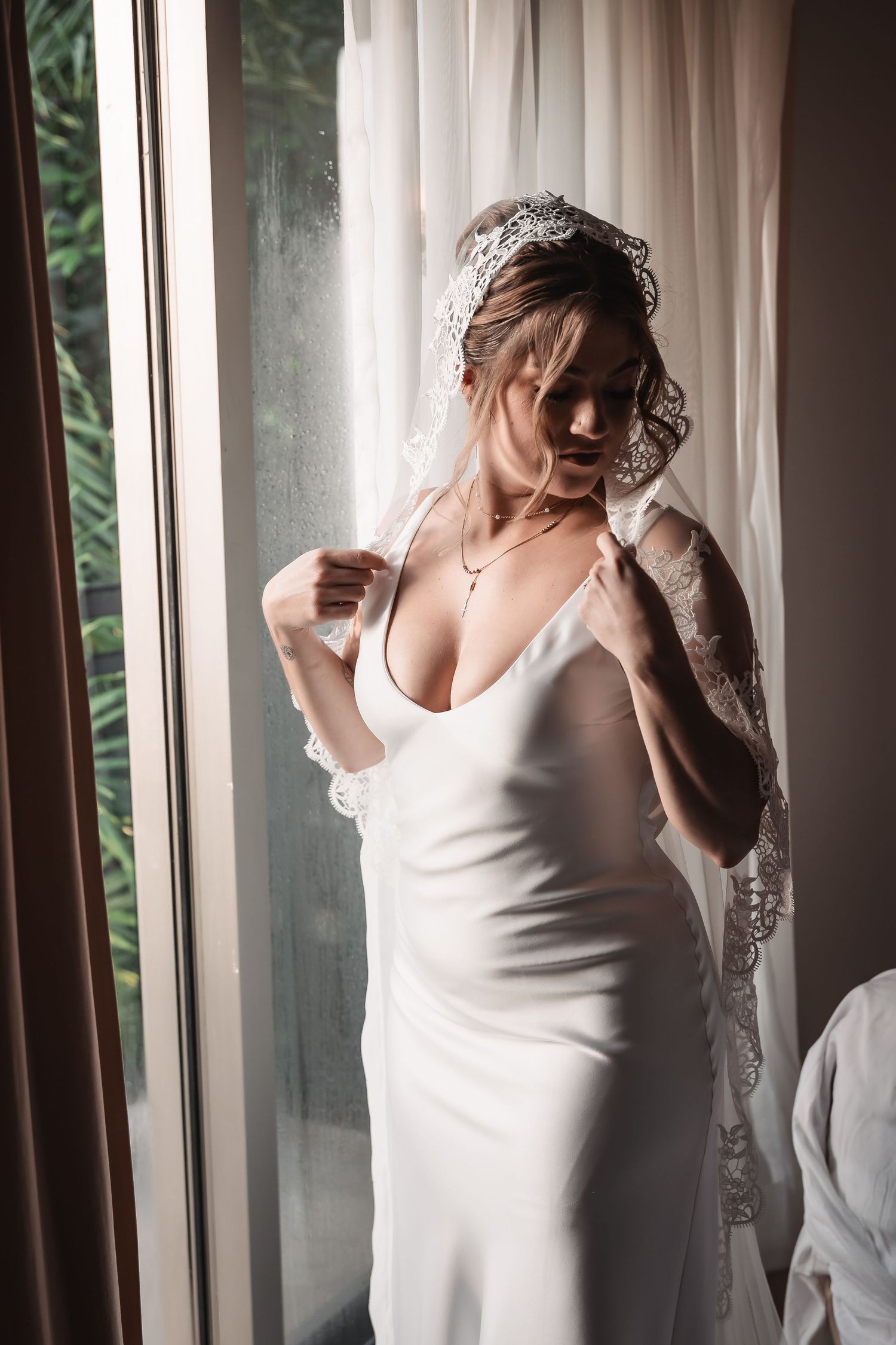 Bride in white slip dress poses by window with soft natural lighting during wedding preparations.