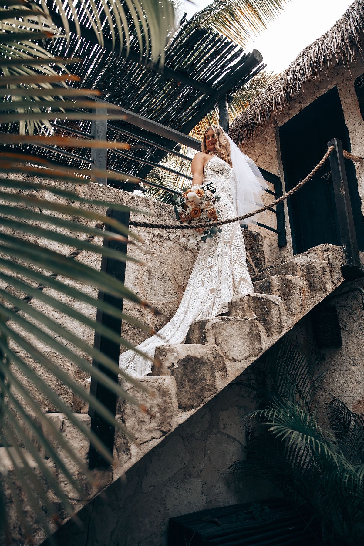 Elegant bridal portraits on stone steps with dramatic palm leaf shadows and thatched roof details.