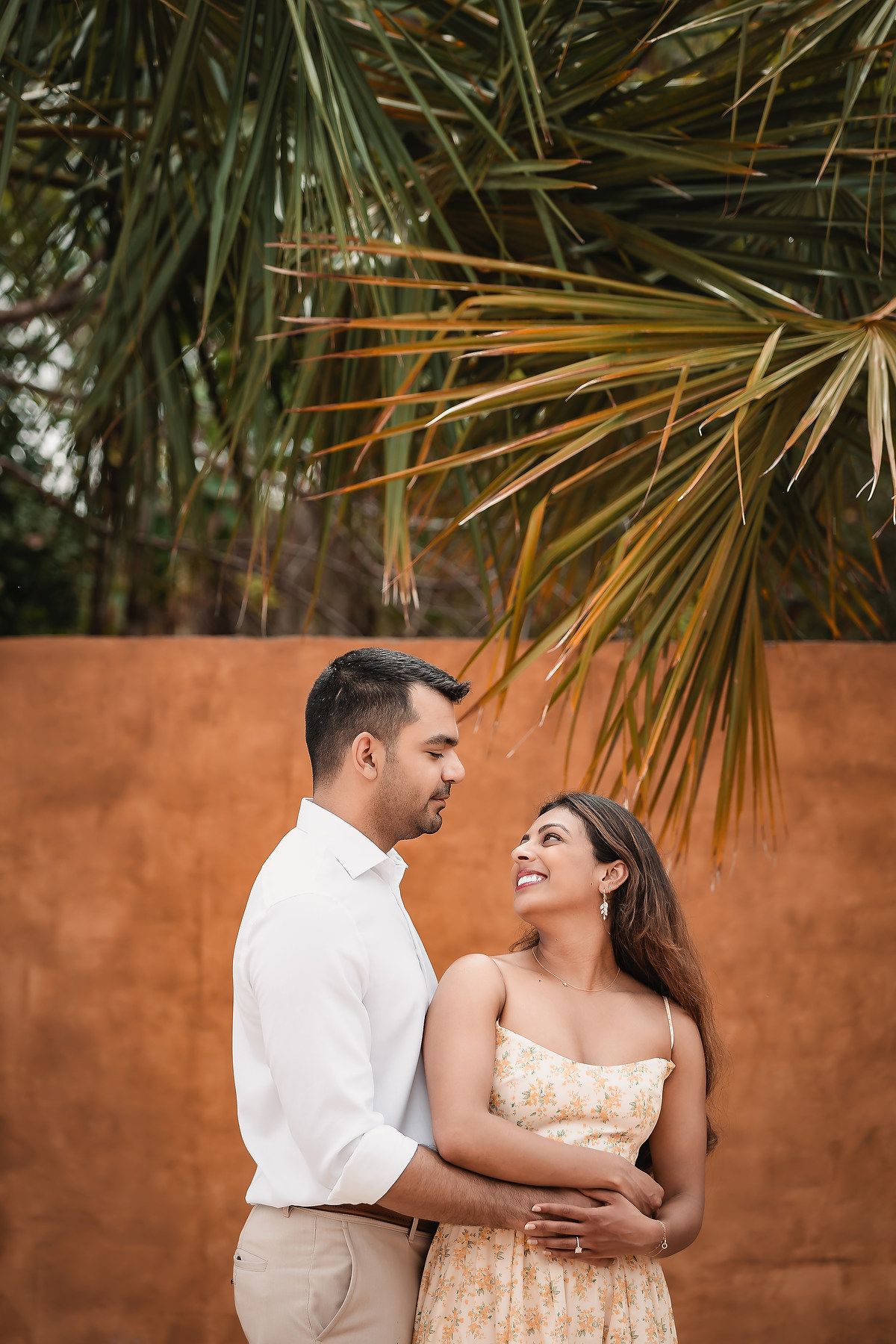 Intimate couple portrait against terracotta wall with tropical plants and palm fronds.