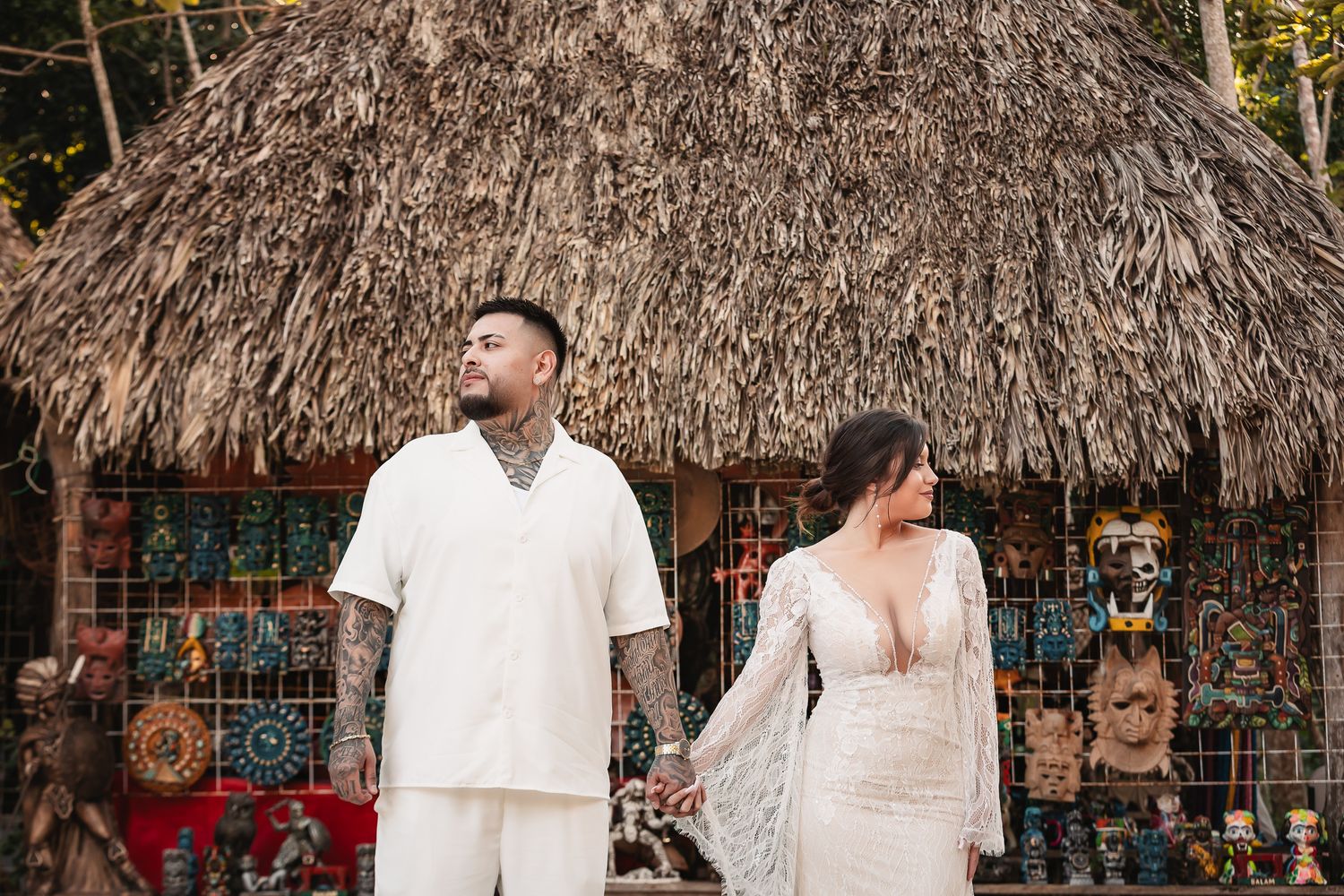 A couple stands together in front of a rustic thatched-roof hut with decorative wall hangings.