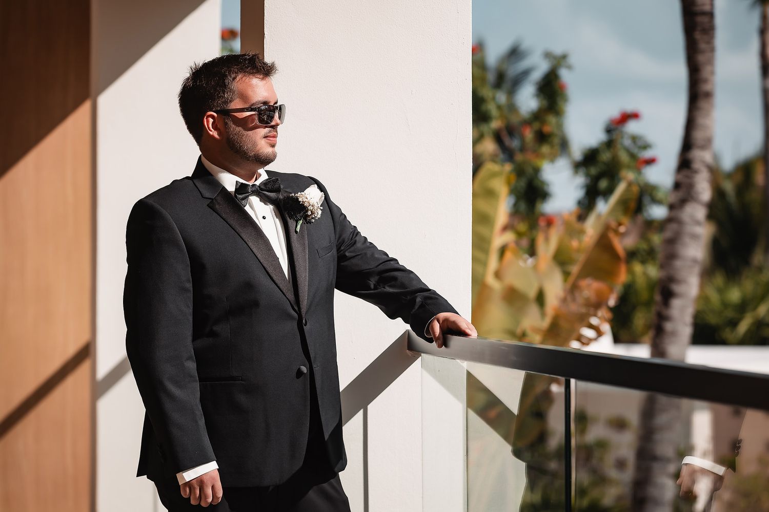 A person in a black tuxedo and sunglasses stands on a balcony with palm trees visible in the background.