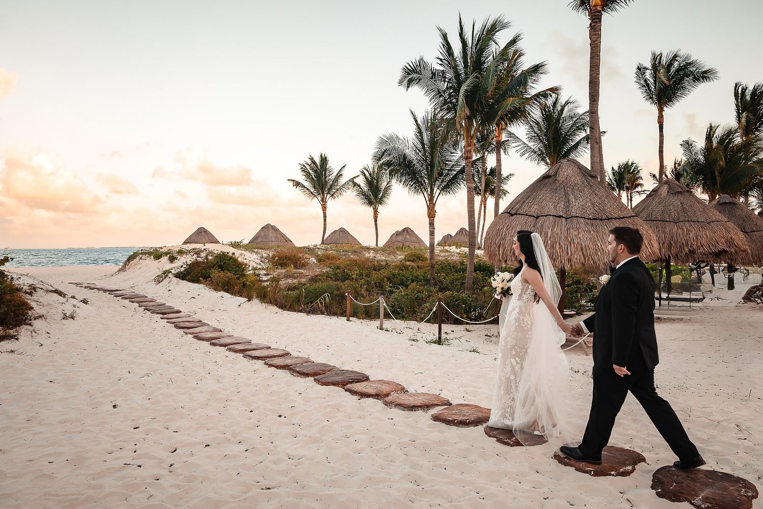 Wedding couple walks along tropical beach with palm trees and thatched umbrellas at sunset.
