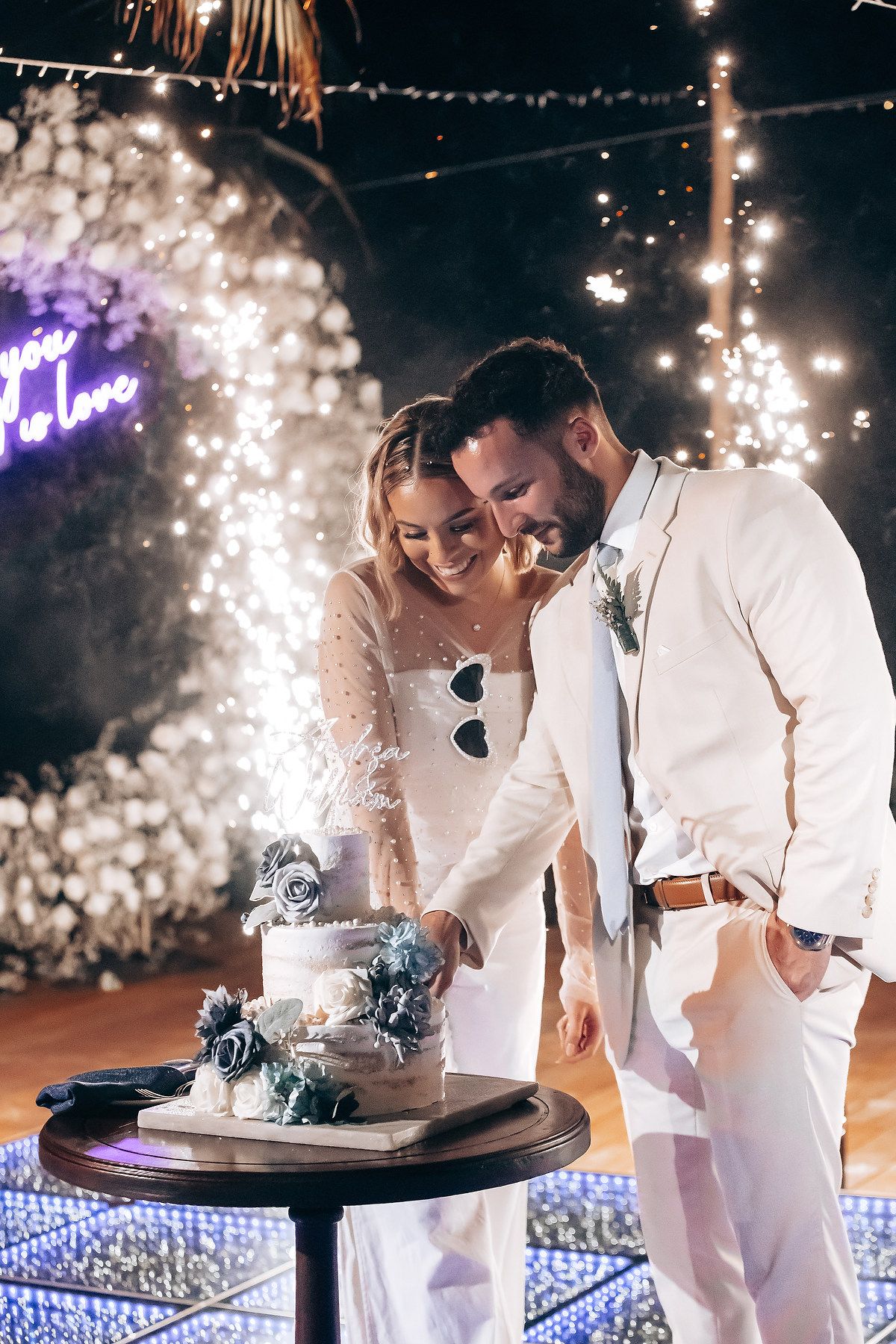 Wedding couple cutting their cake together under romantic string lights at night.