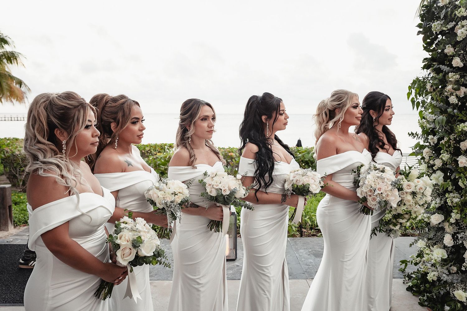 A row of bridesmaids in white dresses holding bouquets pose together at an outdoor wedding.