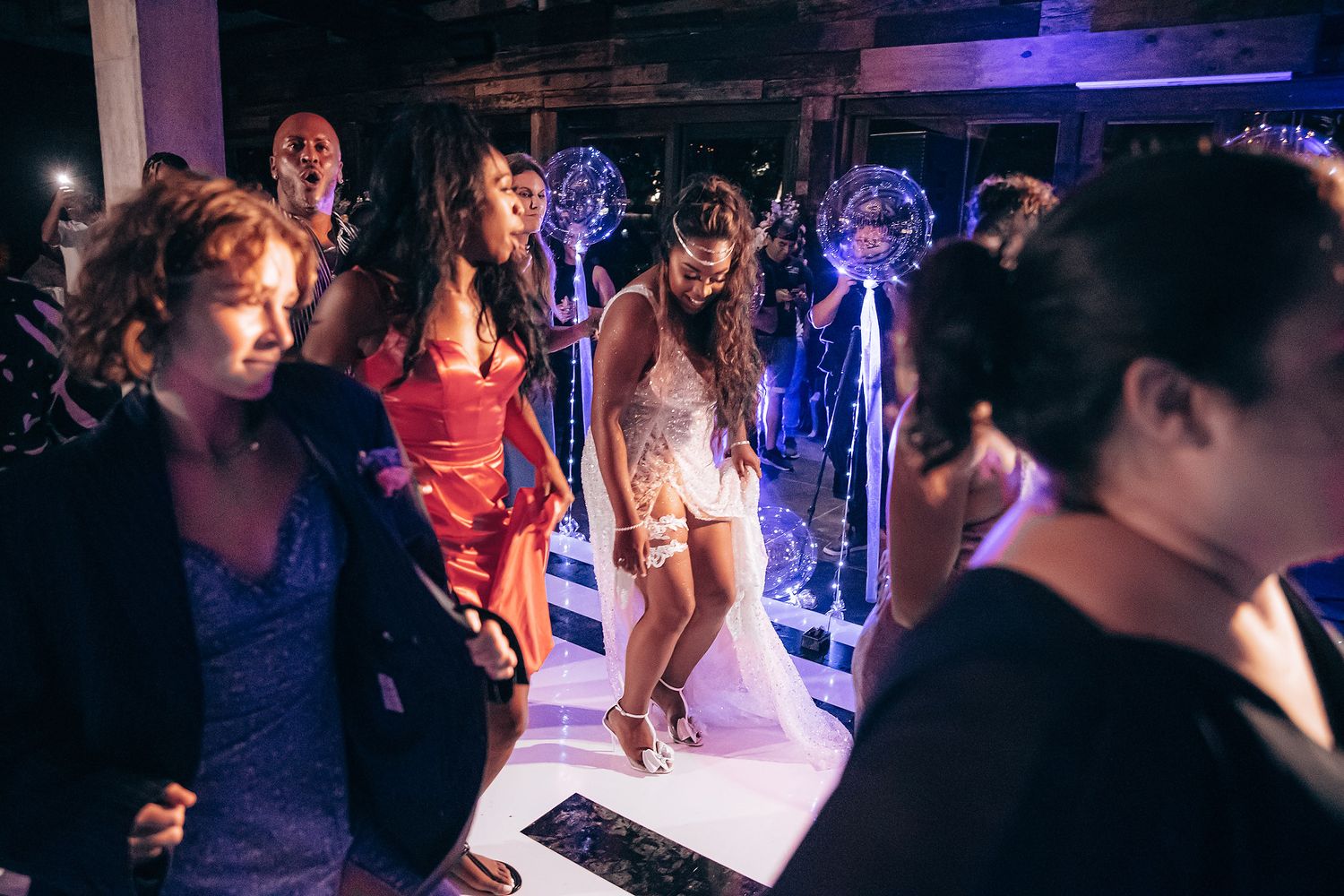 Wedding guests dance and celebrate on a dimly lit dance floor with illuminated decorations in the background.