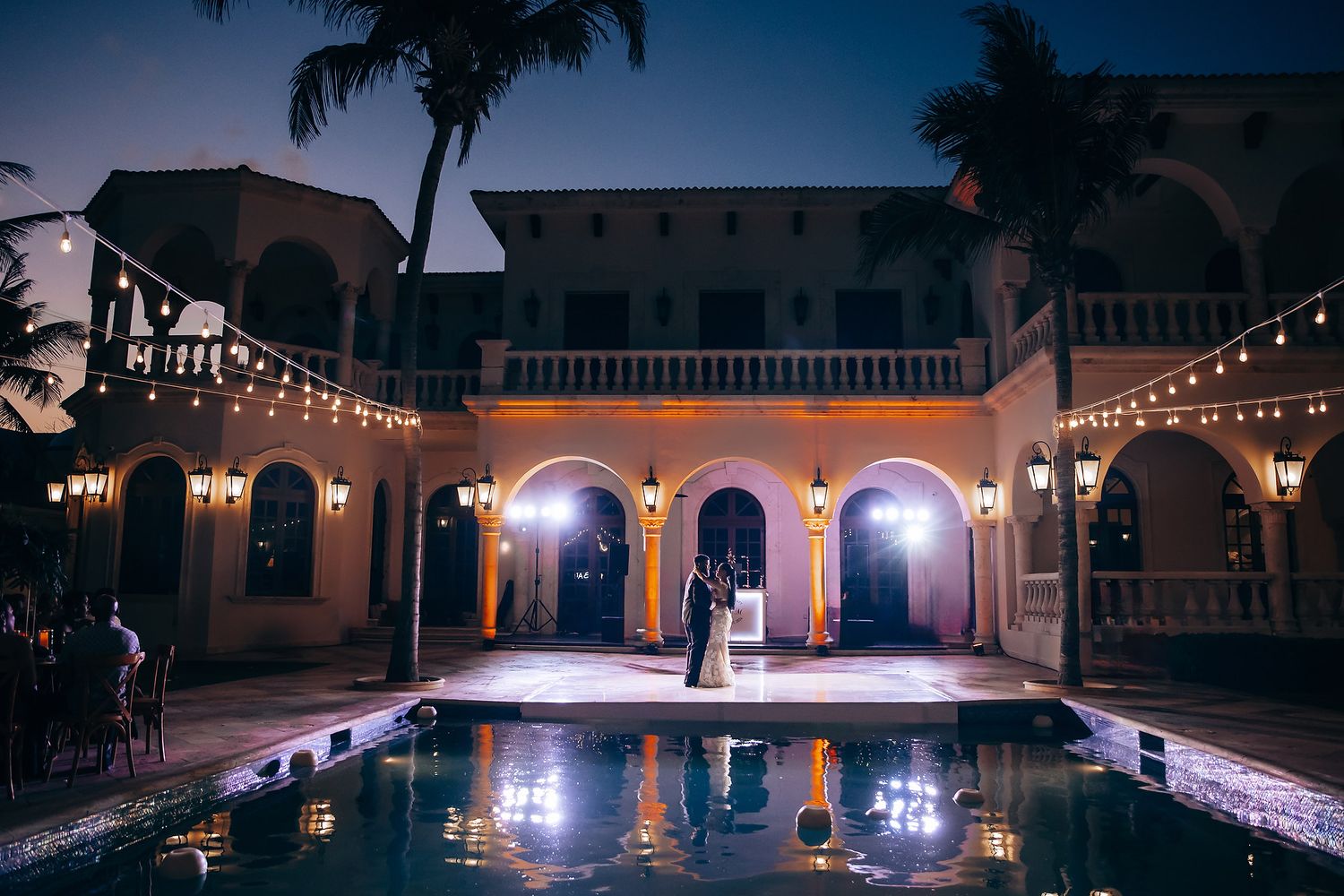 String lights illuminate an outdoor wedding venue at dusk with palm trees in silhouette.