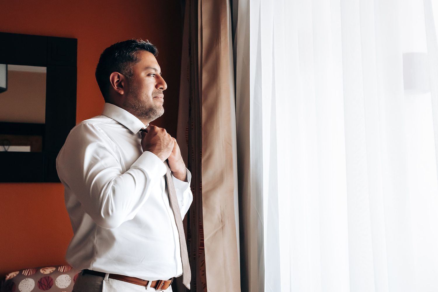 A person in a white shirt stands by a window, looking contemplative in warm lighting.