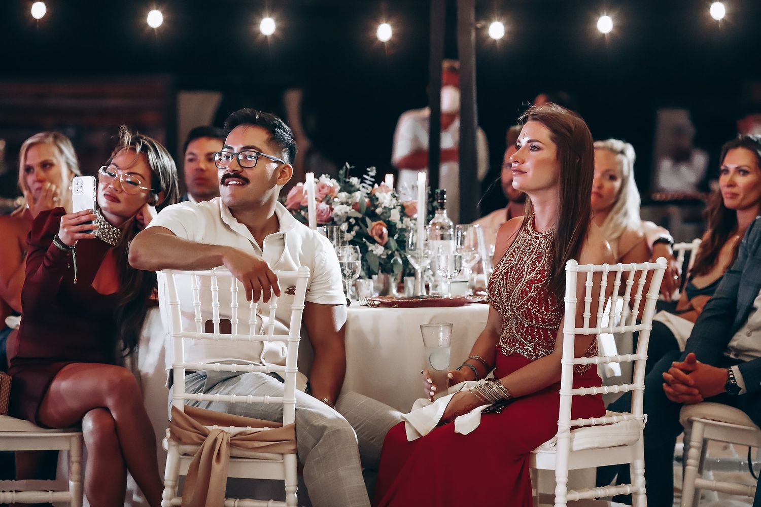 Wedding guests dance and celebrate under string lights at an indoor reception venue.