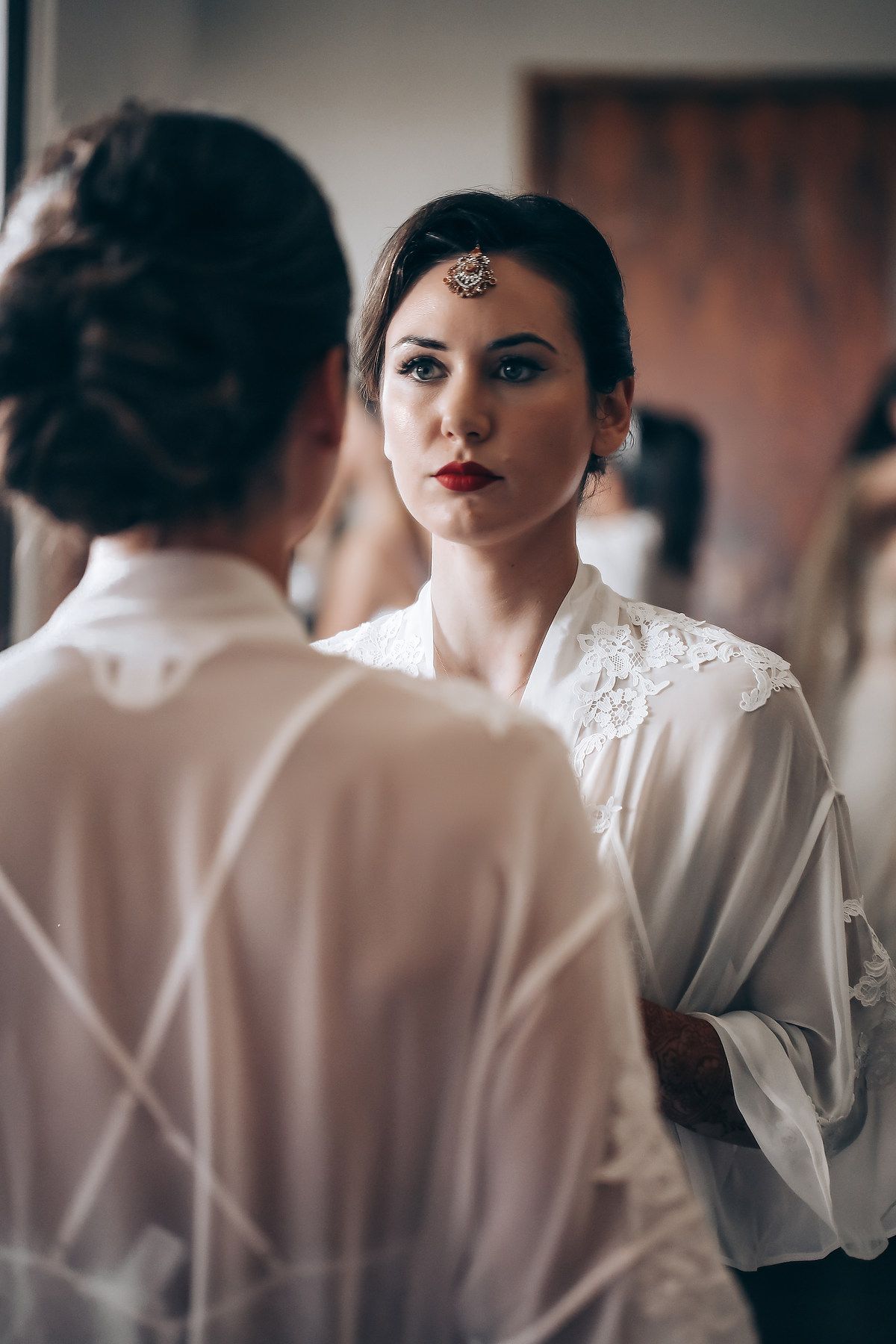 A woman in a white silk robe with traditional Indian bridal jewelry looks at her reflection in a mirror.