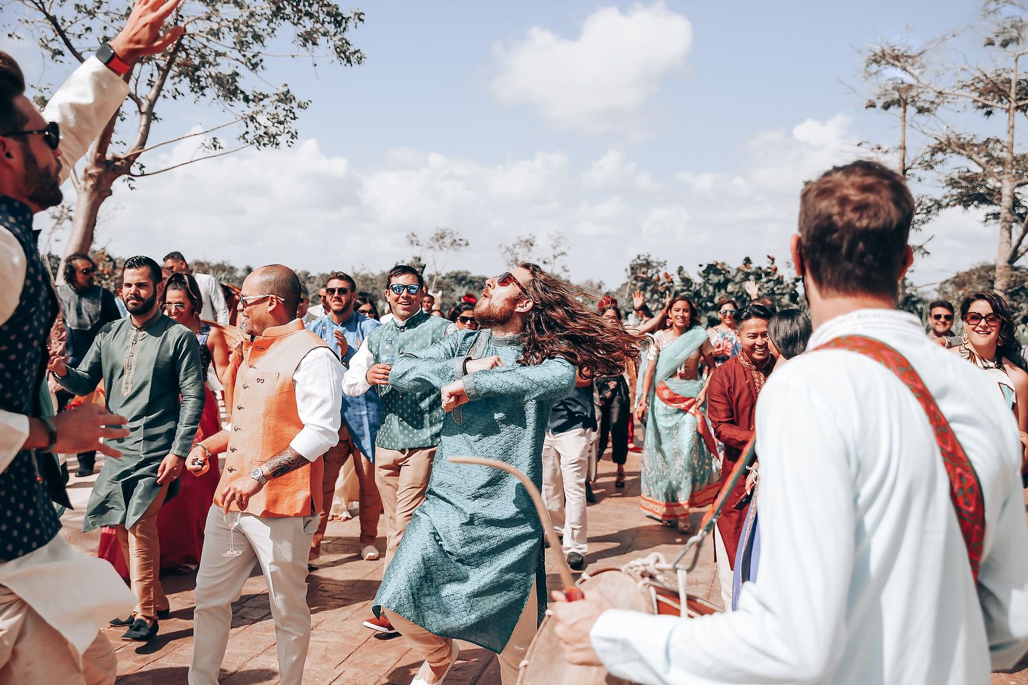 A crowd of festival-goers dancing and celebrating outdoors under a bright blue sky with trees in the background.
