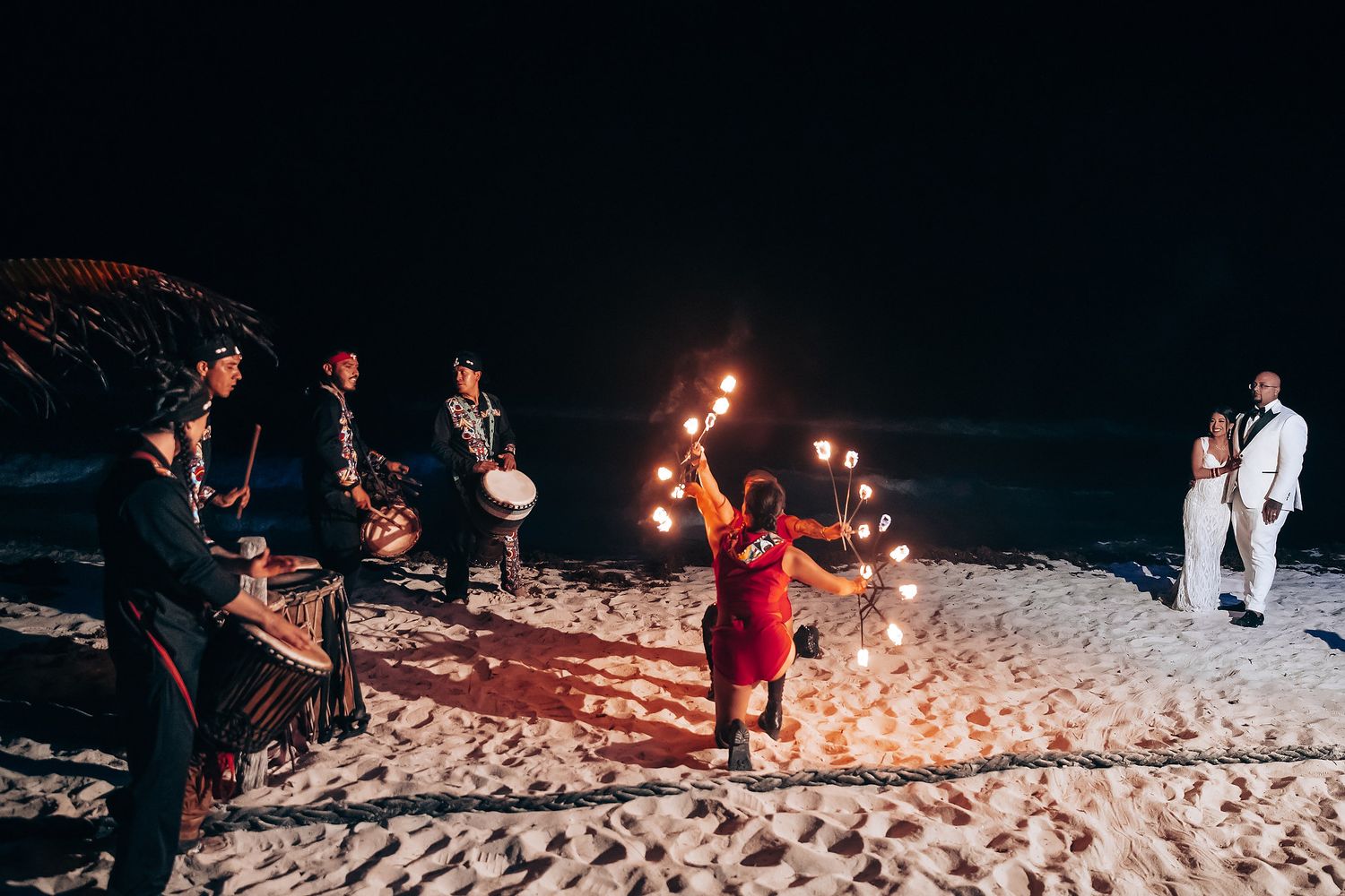 Fire performers entertain wedding guests on a sandy beach during an evening celebration.