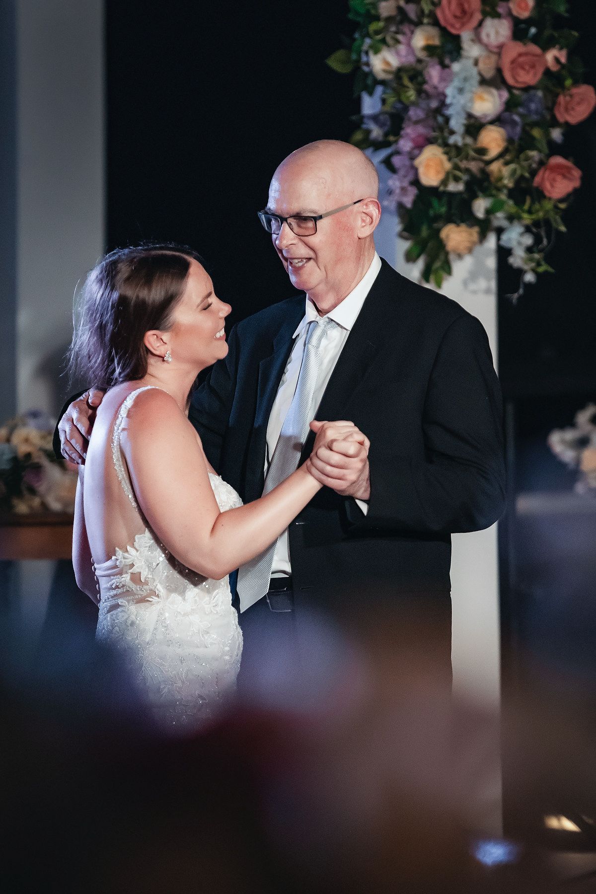 Bride and her father share a joyful first dance at wedding reception with floral arrangement backdrop.