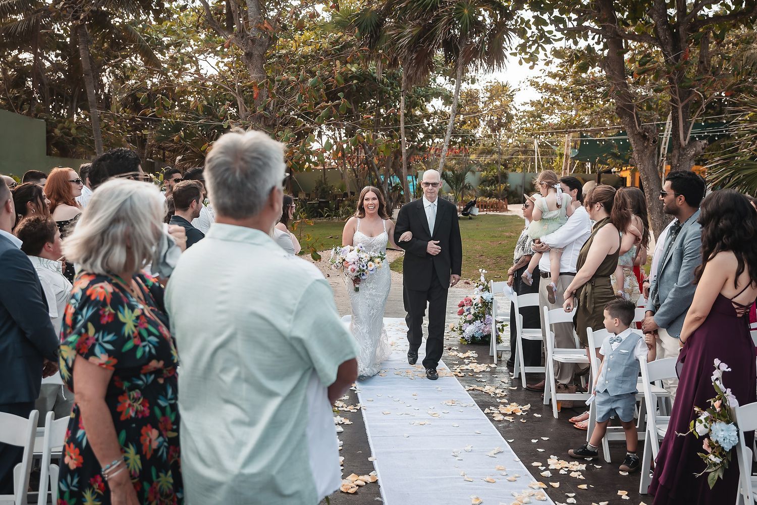 Bride walks down outdoor aisle with escort at garden wedding ceremony, guests standing along white chair-lined path.