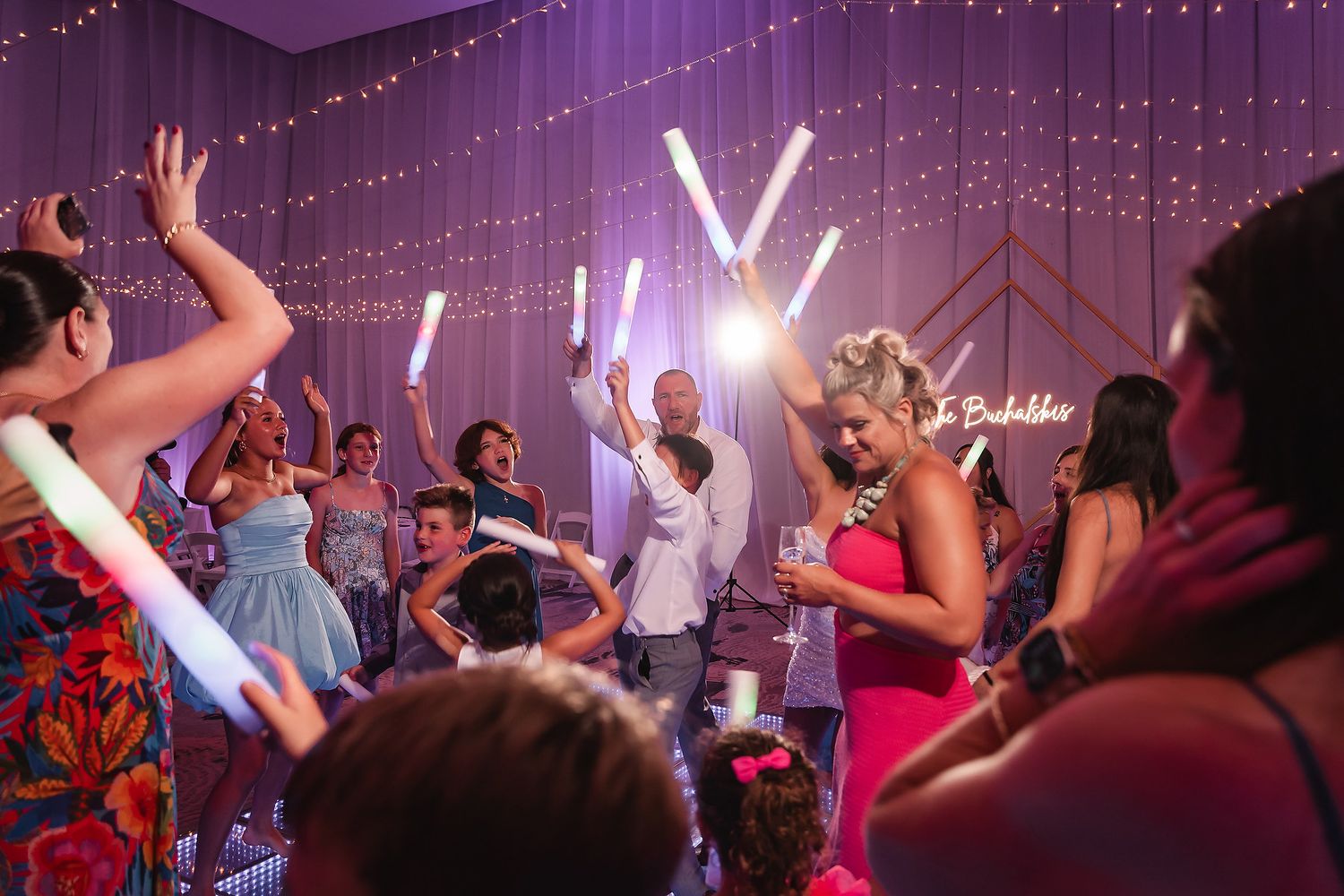 Guests dancing and celebrating at a lively party, holding glowing light sticks under string lights on a purple backdrop.