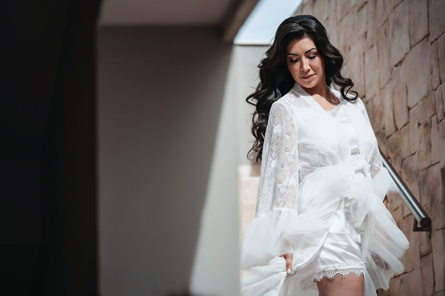 Bride in white lace robe with flowing sleeves stands on balcony, looking down elegantly before wedding ceremony.