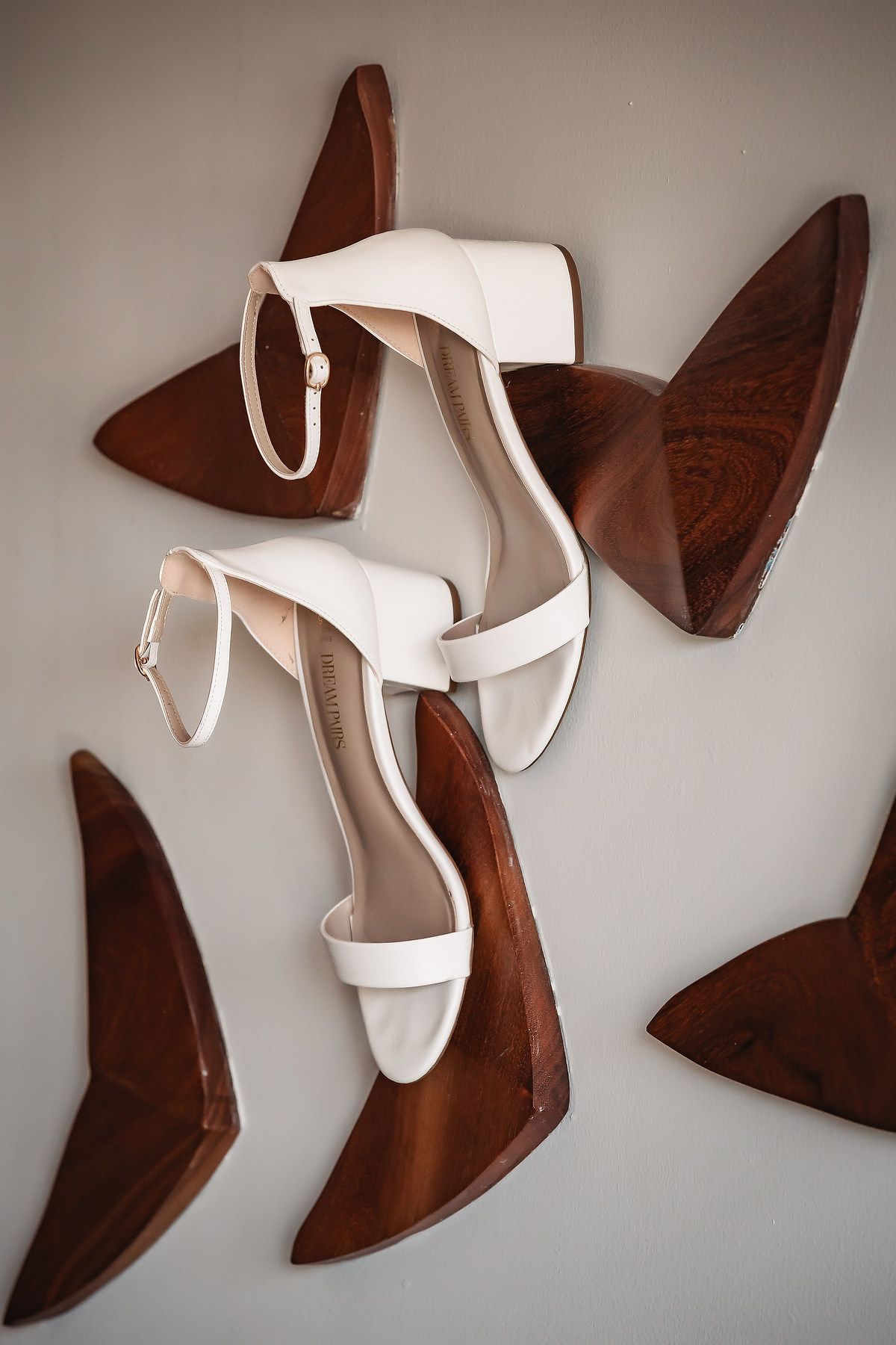 White leather block heel sandals with ankle straps displayed on wooden shoe forms against a gray background.