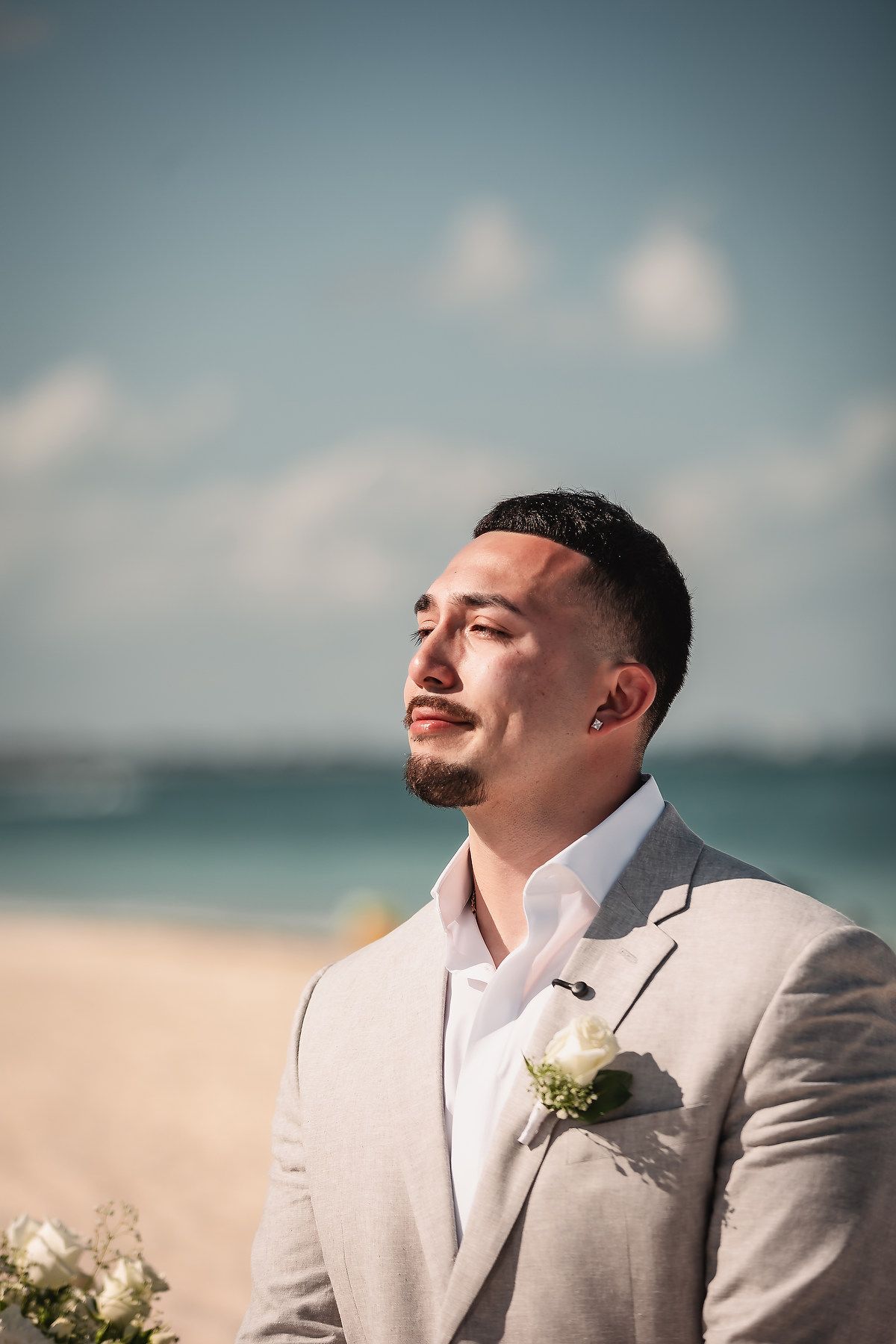 Groom in a light gray suit with a white boutonniere stands on a sunny beach, gazing thoughtfully into the distance.