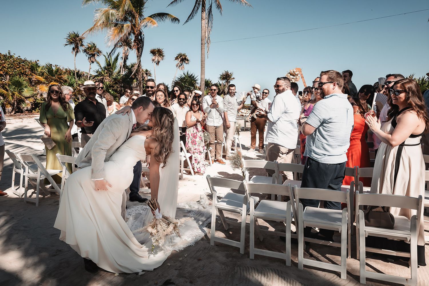 Bride and groom share a romantic kiss during outdoor beach wedding ceremony with guests watching.
