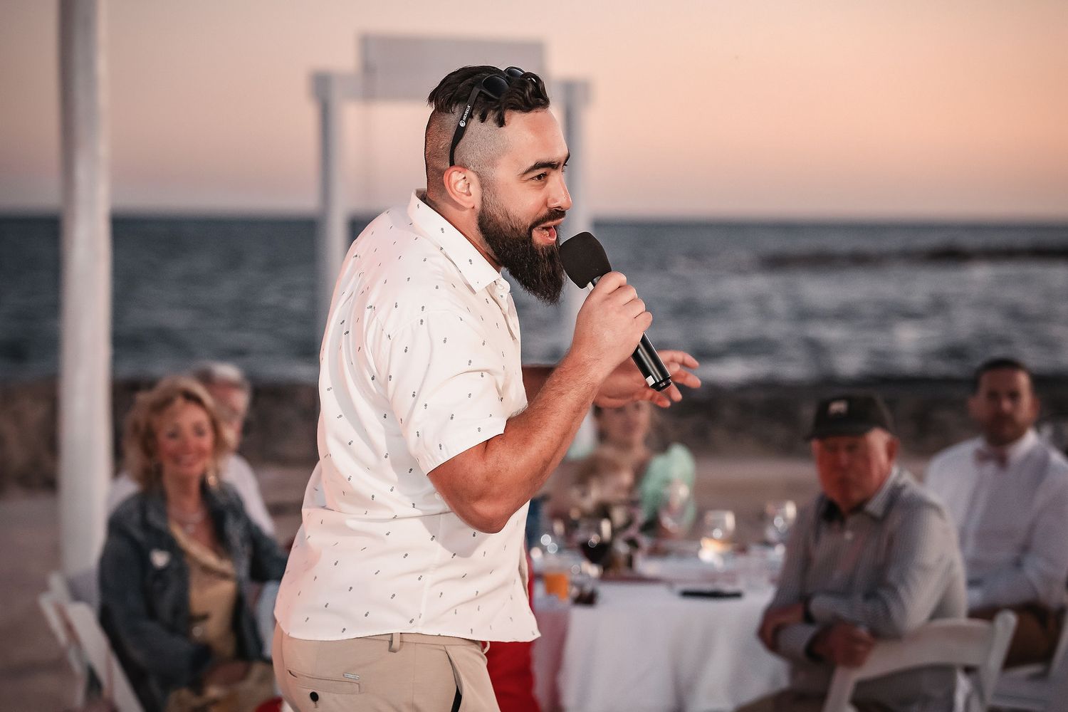 Man giving a speech at an outdoor beach wedding reception at dusk, holding a microphone.