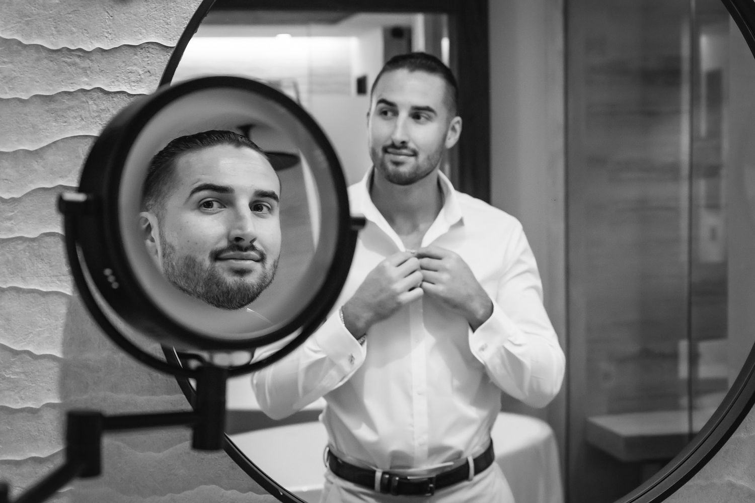 Black and white photo of a man in white shirt adjusting collar, reflected in round mirror.