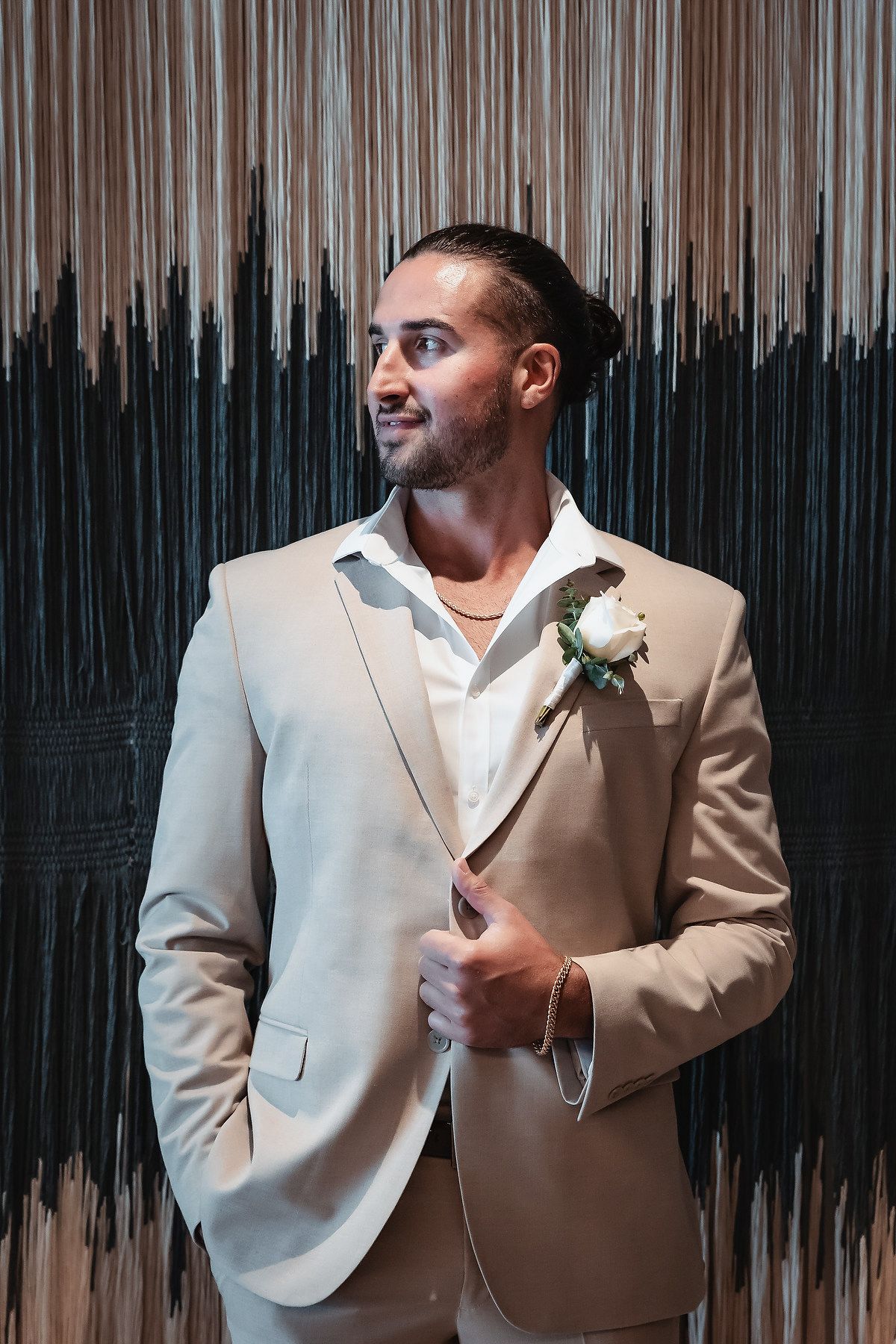 Groom in beige suit with white boutonniere poses confidently against decorative backdrop at wedding.