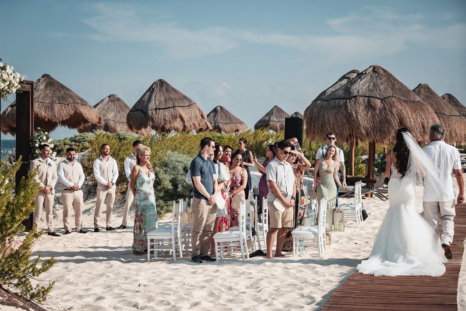 Beach wedding ceremony with guests seated on white chairs, thatched palapas, and a bride in a white gown on a sandy shore.