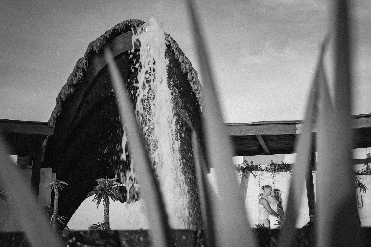 Black and white photo of a couple embracing near a circular waterfall feature, framed by tropical plants.