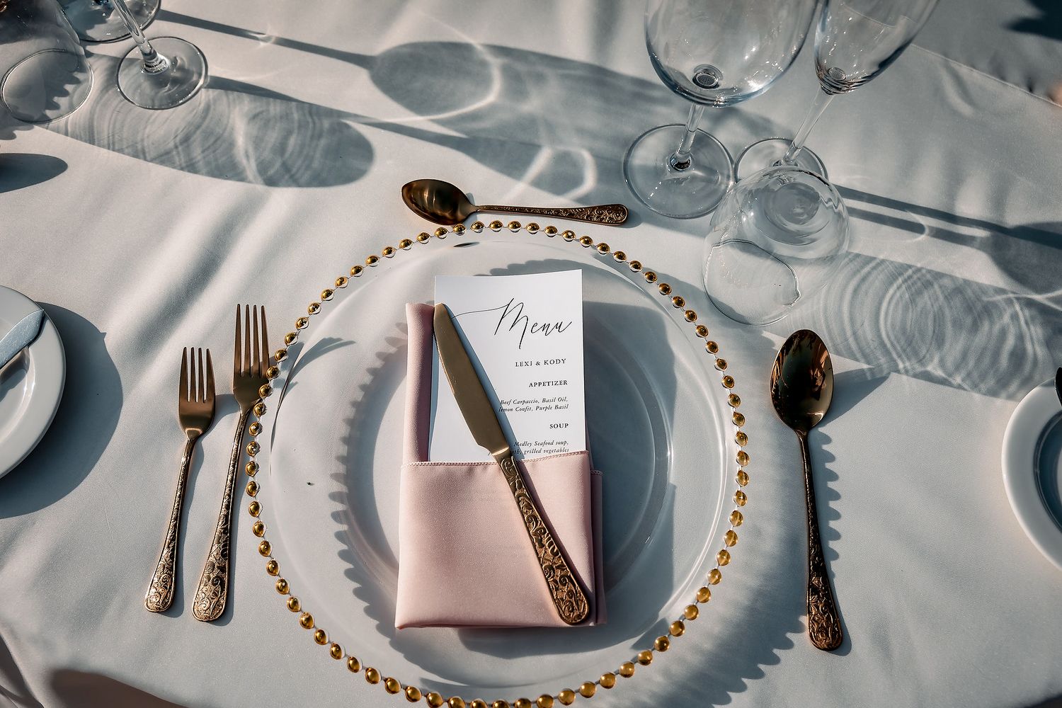 Elegant wedding place setting with gold beaded charger, pink napkin, menu card, and gold cutlery on white tablecloth.