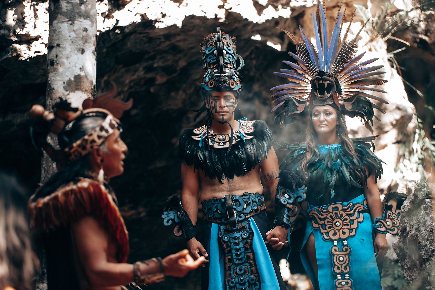 Three performers in elaborate Mayan ceremonial costumes with feathered headdresses and blue garments in a jungle setting.