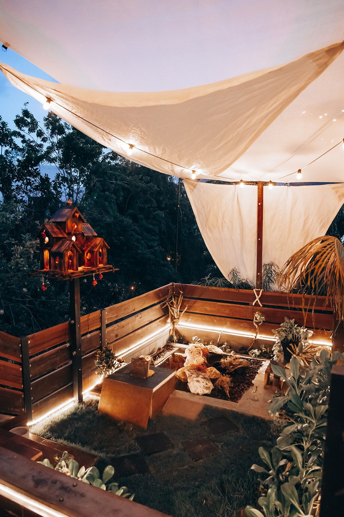 Cozy outdoor patio with string lights, sail shade canopy, wooden benches, fire pit, and decorative birdhouse at dusk.