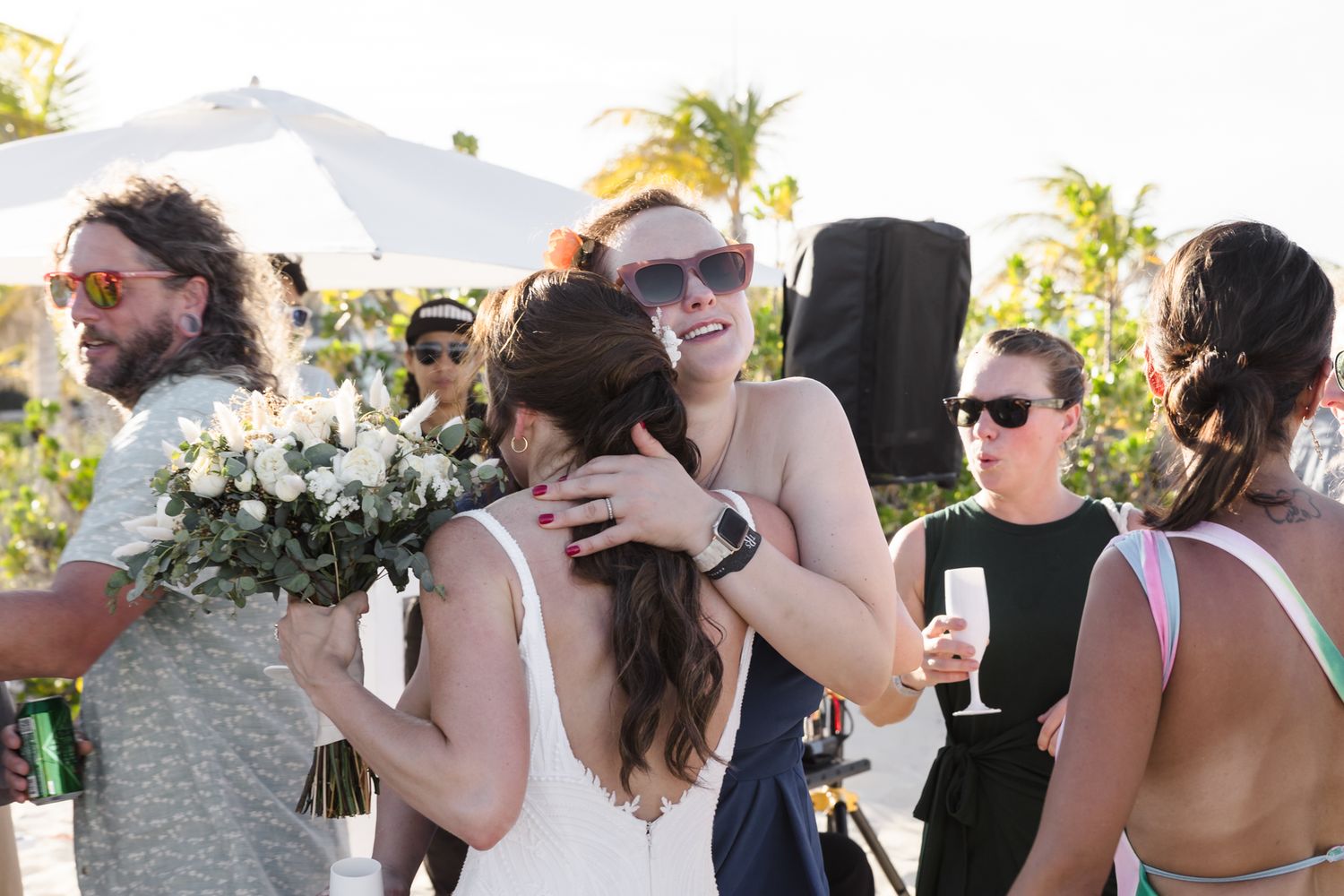 Guests hugging and celebrating at an outdoor beach wedding reception with bouquet and champagne.