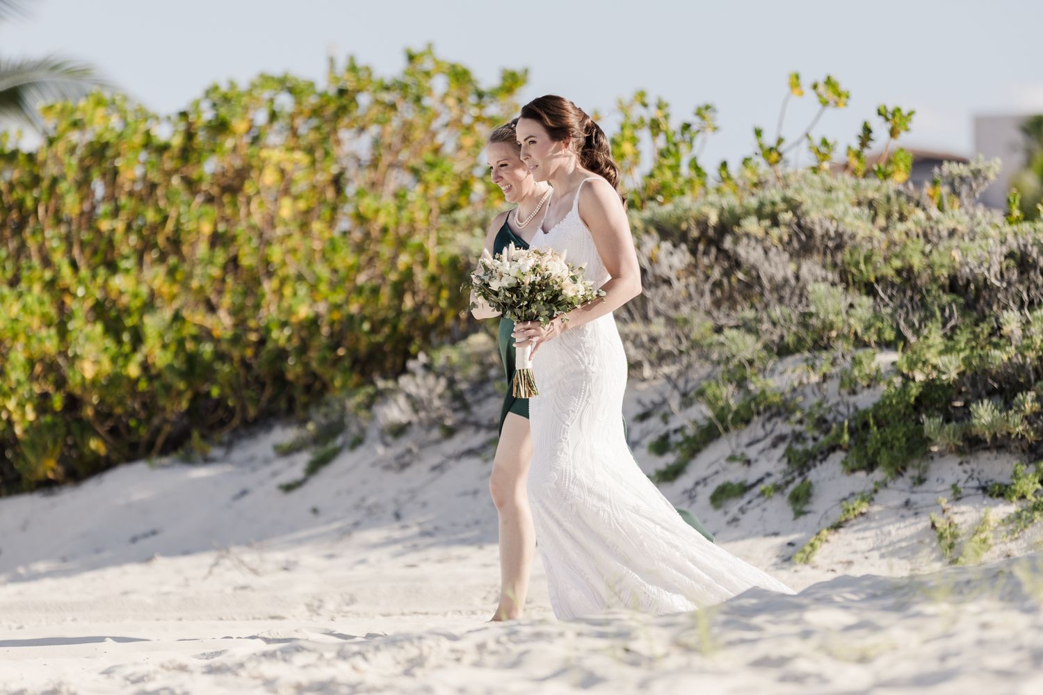Bride in white gown holding bouquet walks on sandy beach with groom, lush greenery in background.