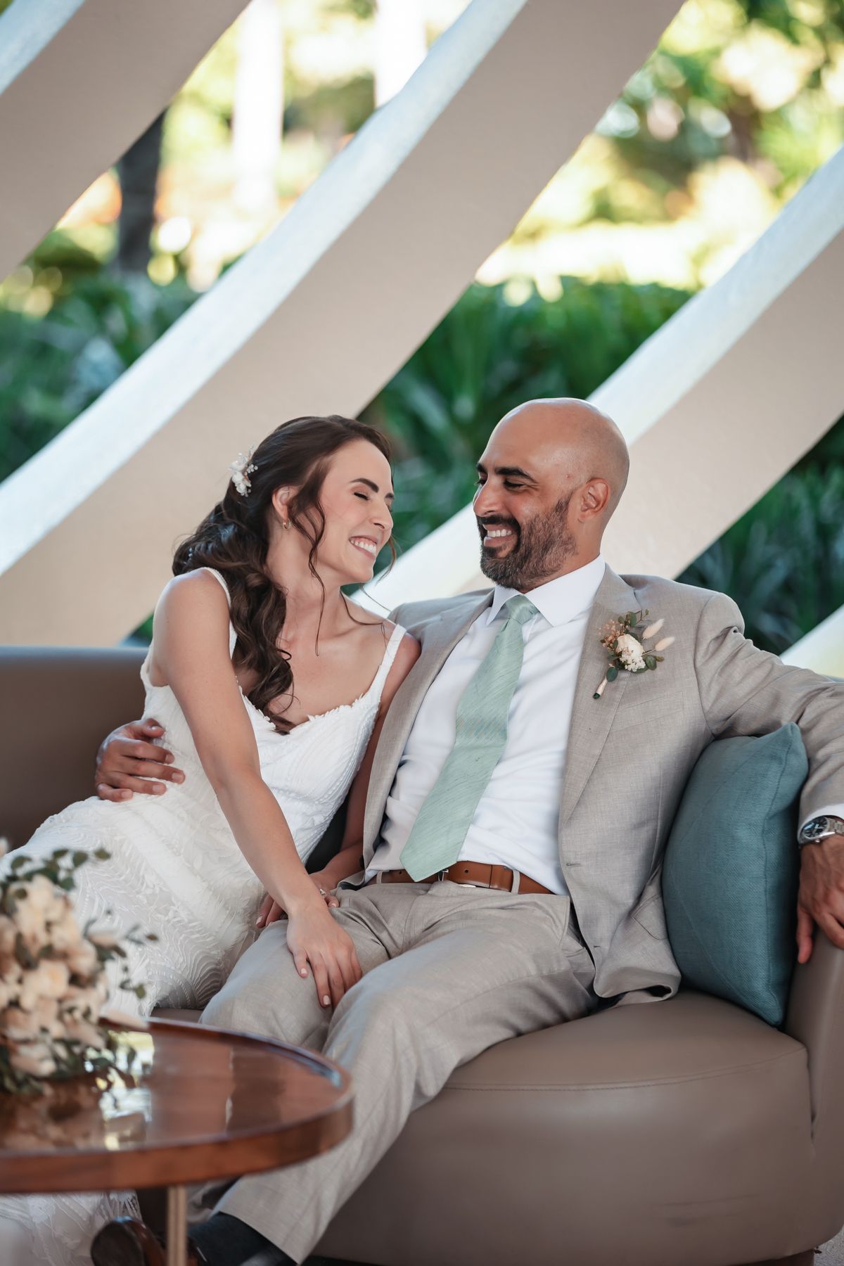 Bride and groom sitting together on a couch, smiling at each other, groom in gray suit with green tie, bride in white dress.