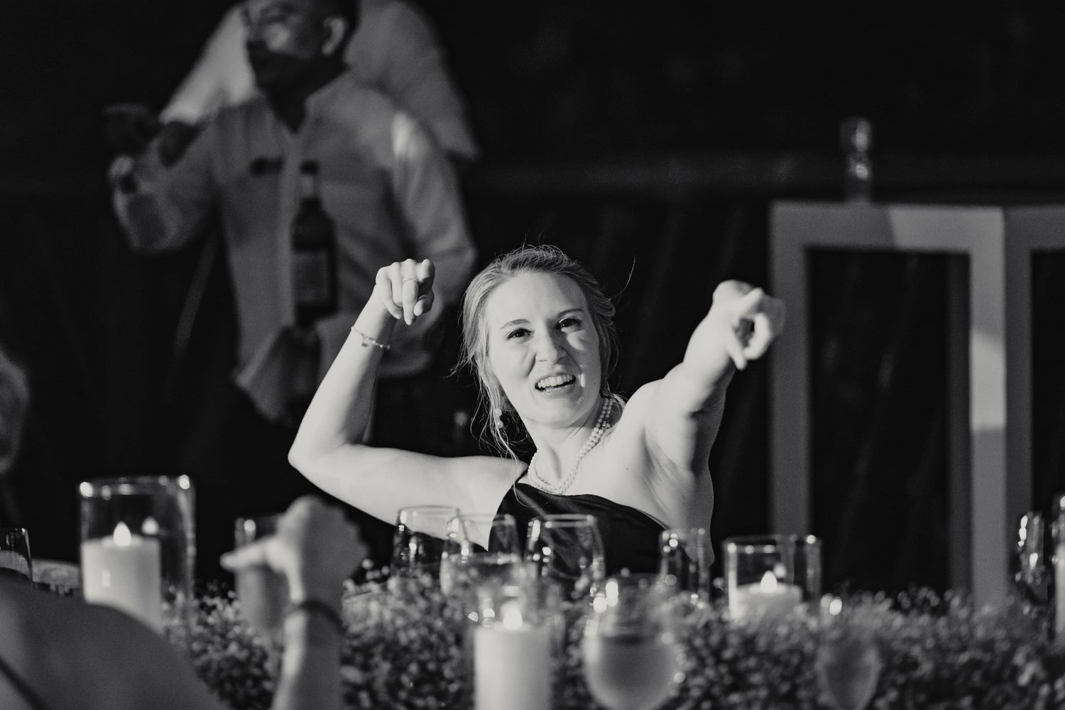 Black and white photo of a joyful bride celebrating at her wedding reception table with raised fists.