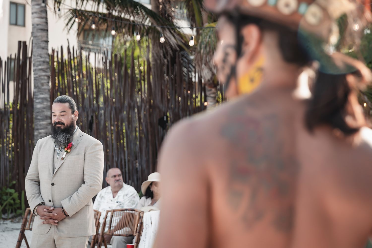 Groom in beige suit watches tattooed shirtless officiant during outdoor beach wedding ceremony in Mexico.