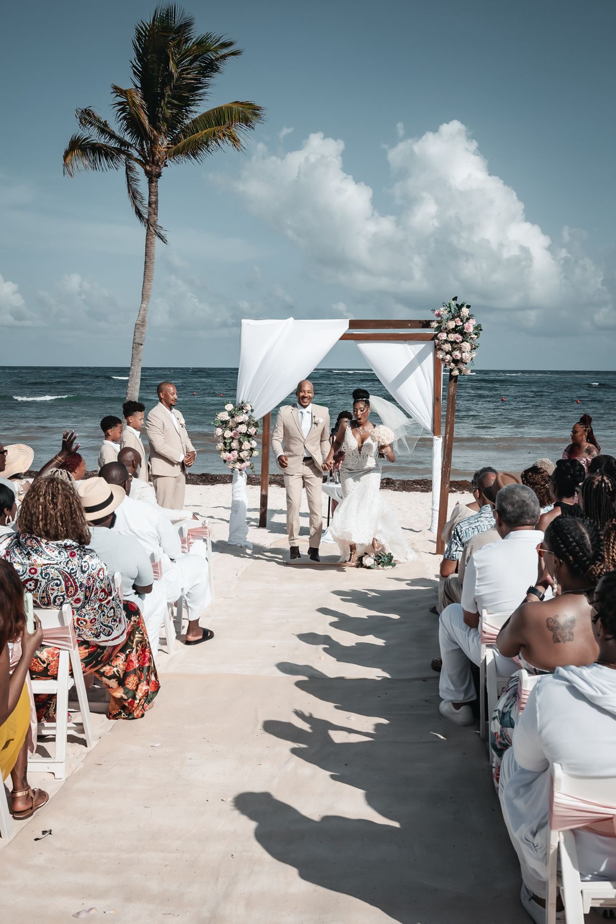 Outdoor beach wedding ceremony with couple at floral arch, guests seated on white chairs, palm tree and ocean in background.