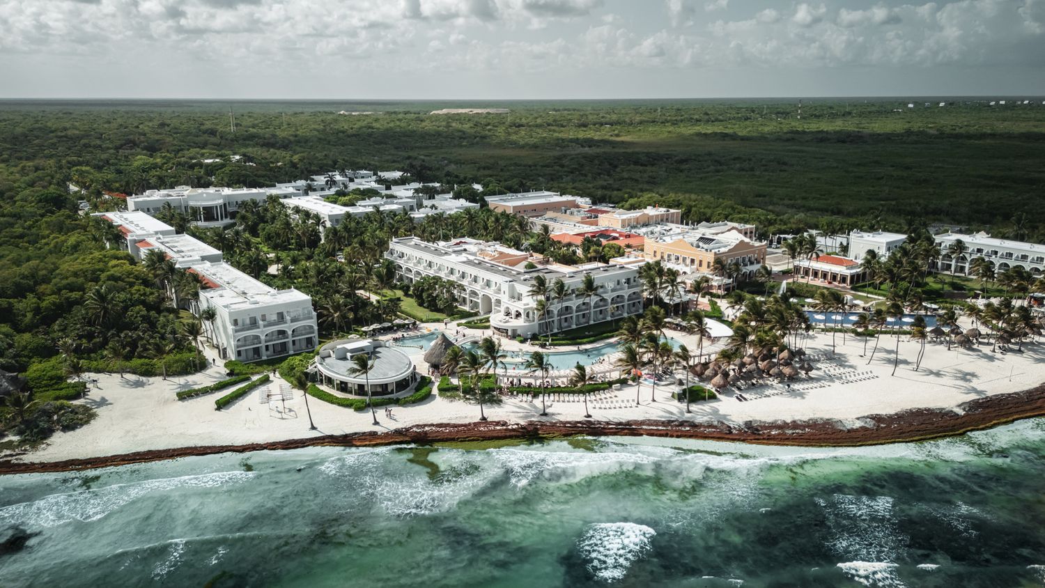 Aerial view of a beachfront resort with white buildings, pool, and lush tropical greenery along a turquoise Caribbean coastline.